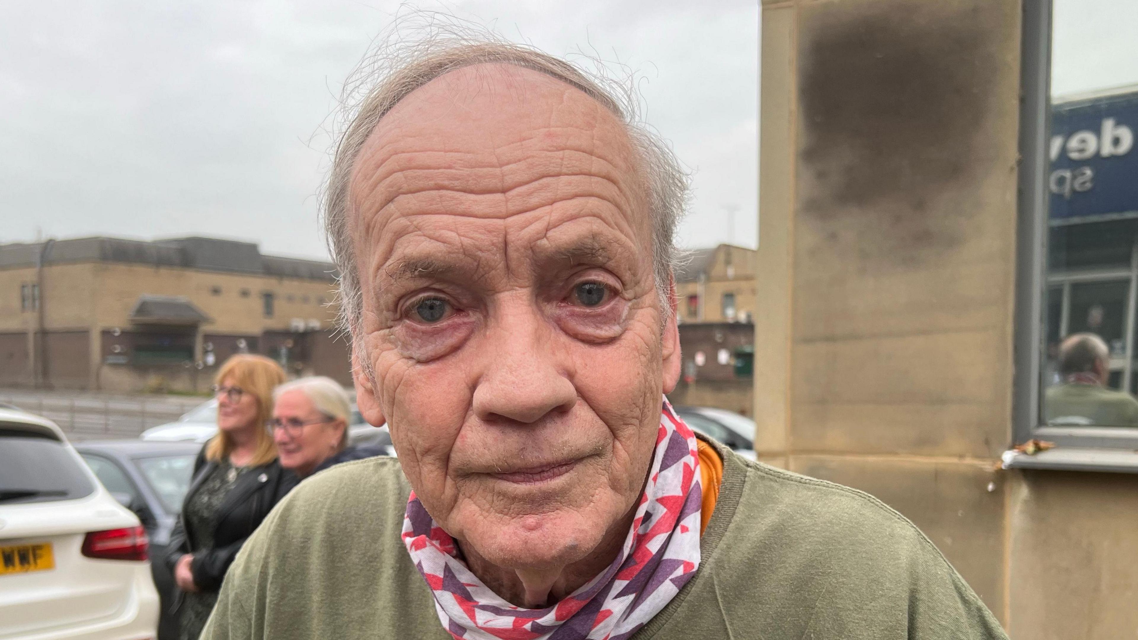 Image shows an older man with a green t shirt and patterned scarf. 
He is standing in front of brown concrete buildings in a car park. 