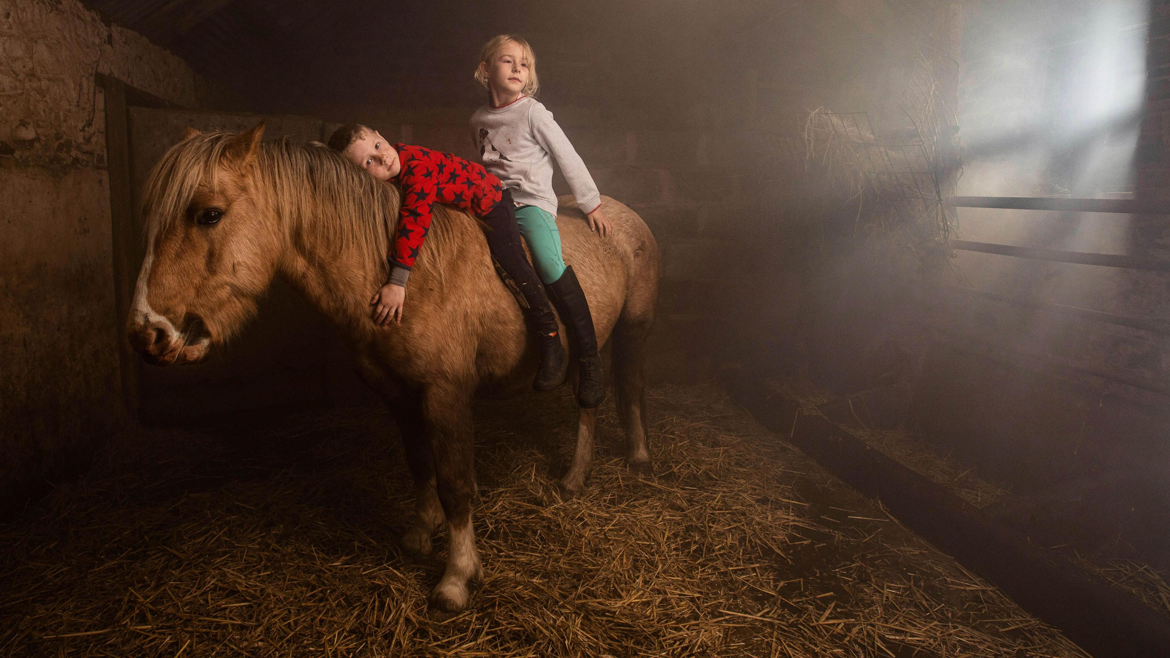 A boy and a girl are on a horse in a stable. The brown horse stands on hay. The boy cuddles the horse, he has black jodhpurs on and a red top with black stars. The girl is behind him and has turquoise jodhpurs with a white top. She has short blonde hair.