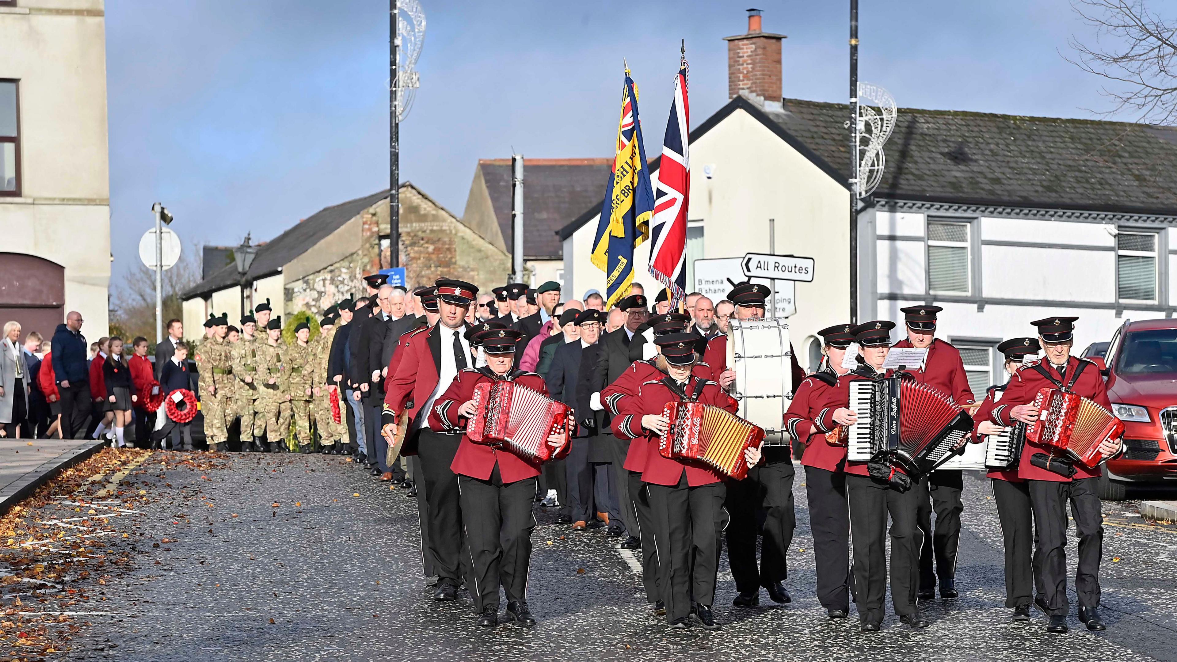 A parade is walking through a road. The people in front are holding instruments. and wearing maroon jackets with back trousers. The union flag is being held by someone in the parade and some people are holding poppy wreaths. 