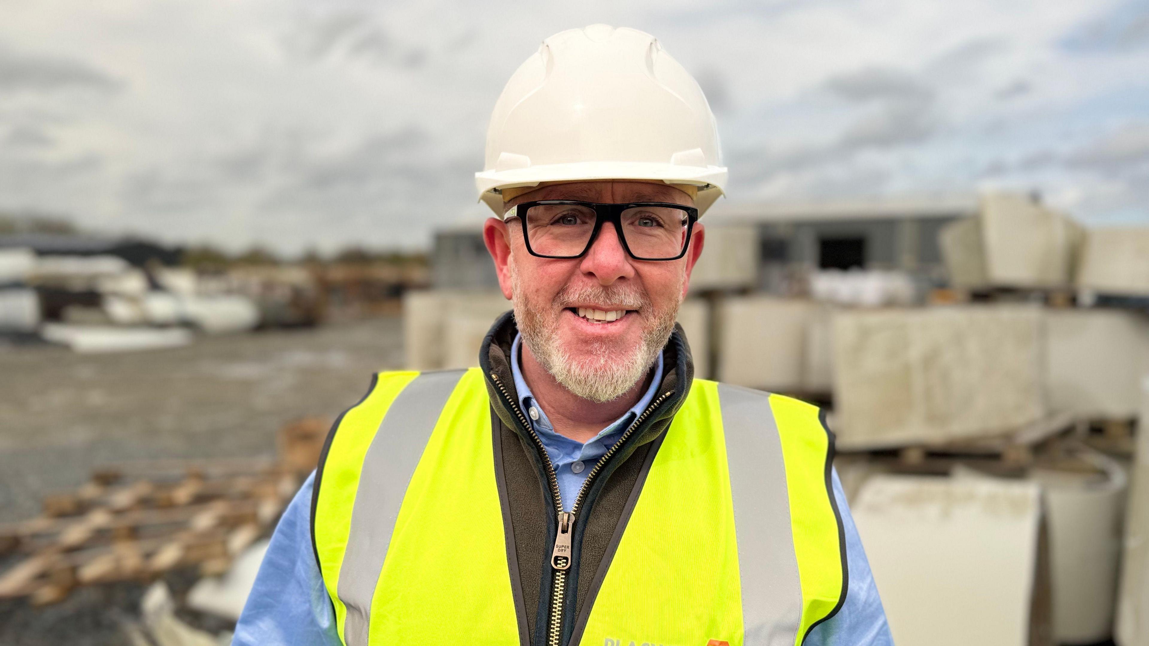 Kieran Kelly, a man with black square-framed glasses and a grey beard, smiles at the camera in an industrial yard.  He is wearing a white helmet, a yellow high-viz vest, a blue shirt and a brown body warmer. 