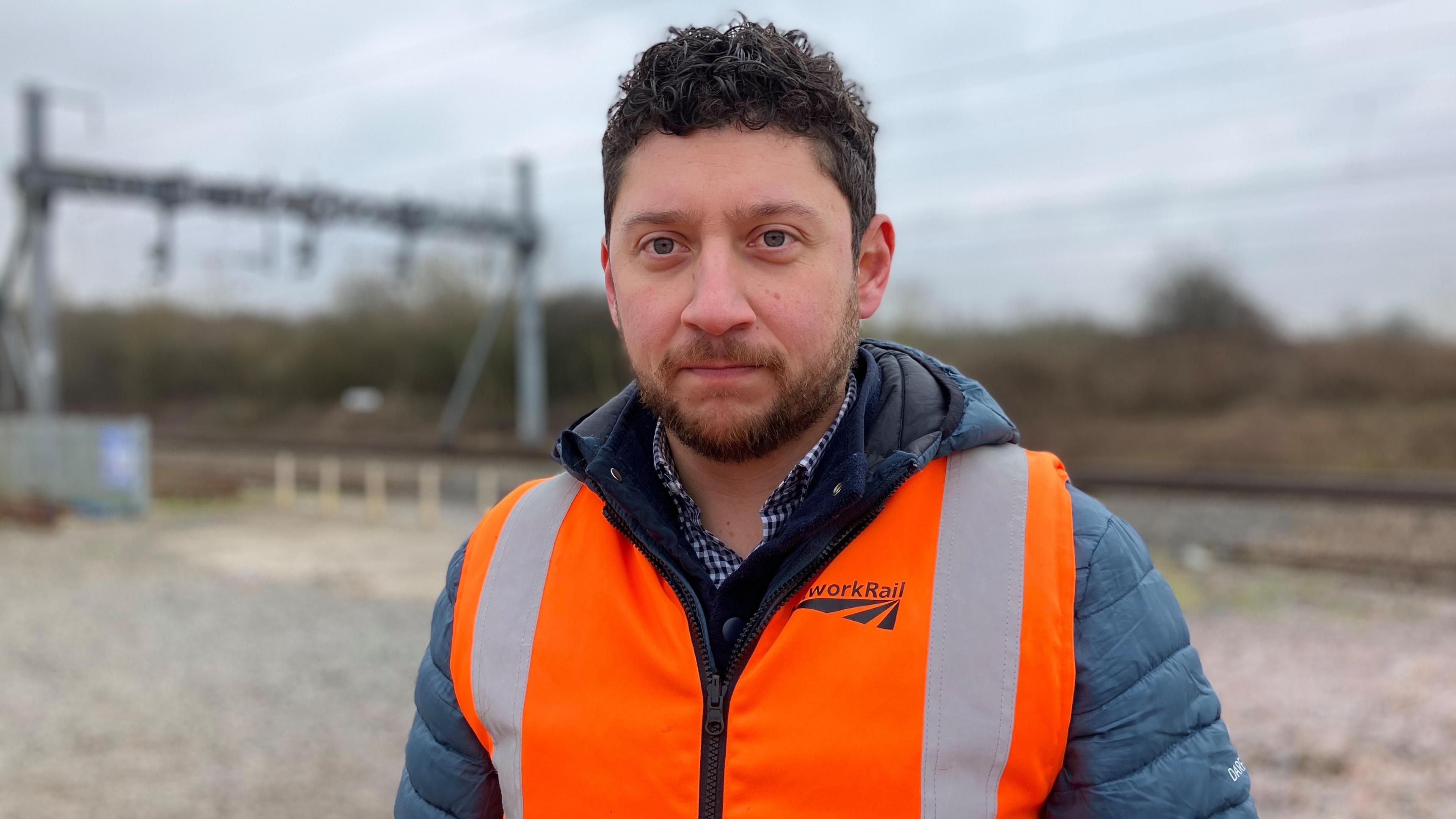 A man with curly brown hair and a light beard stood outside with a railway line and overhead cables behind him. He is wearing an orange high-vis Network Rail jacket.