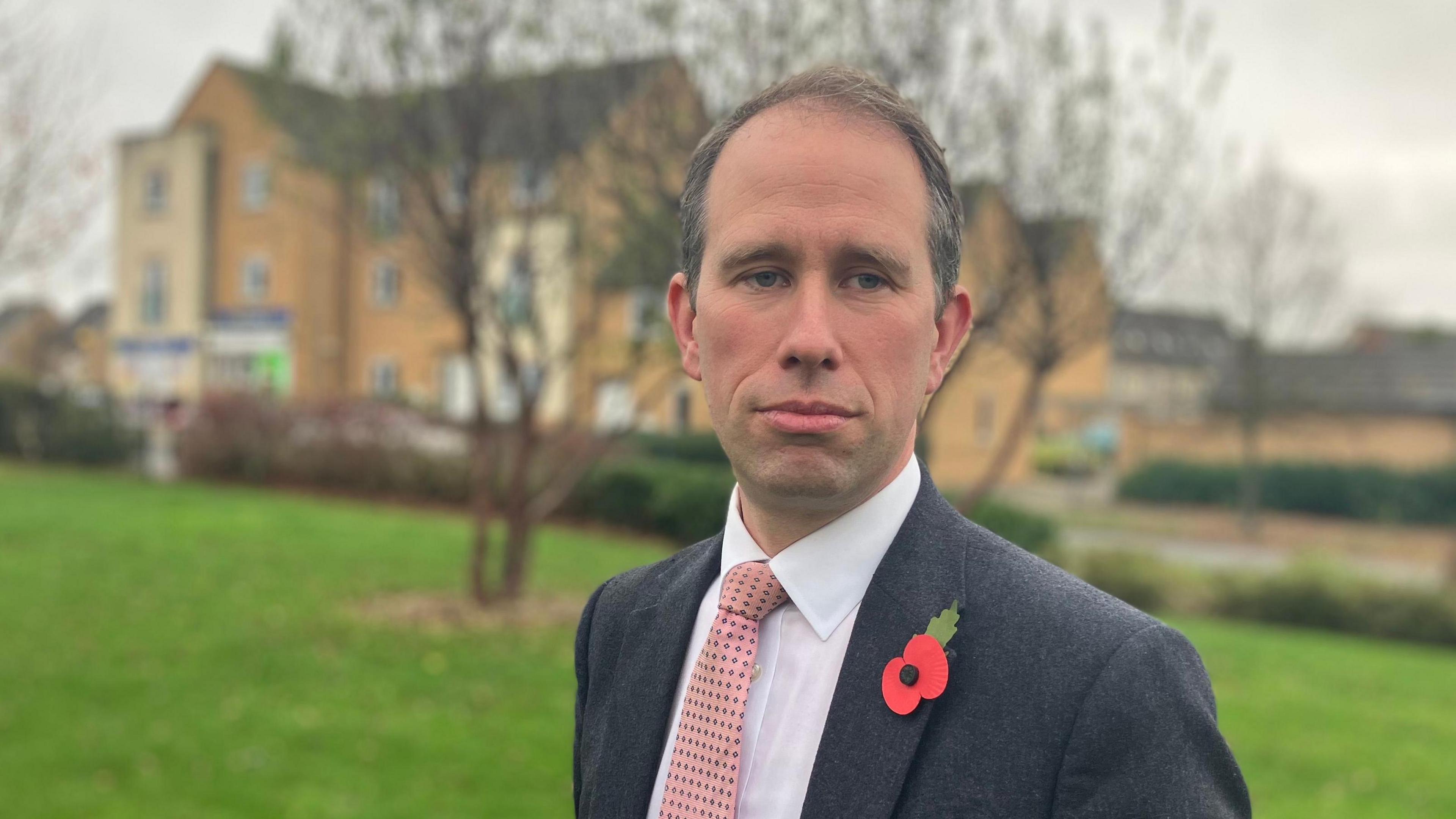 Matthew Barber has short brown hair and is wearing a grey blazer, white shirt and pink tie. A yellow-ish building and a grass field are out of focus behind him.