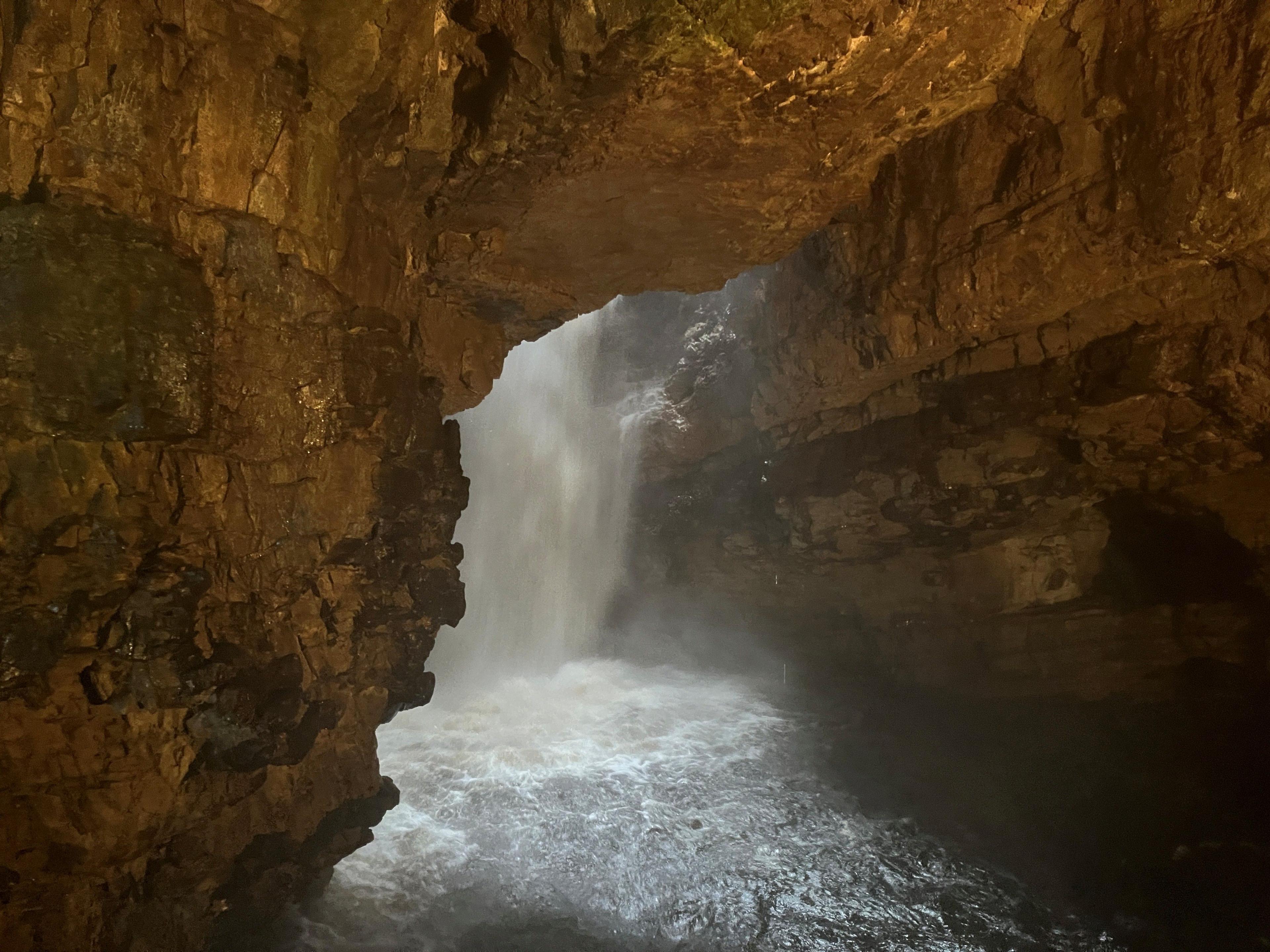 Inside of a cave with a waterfall flowing strongly in the centre. The cave is slightly lit.