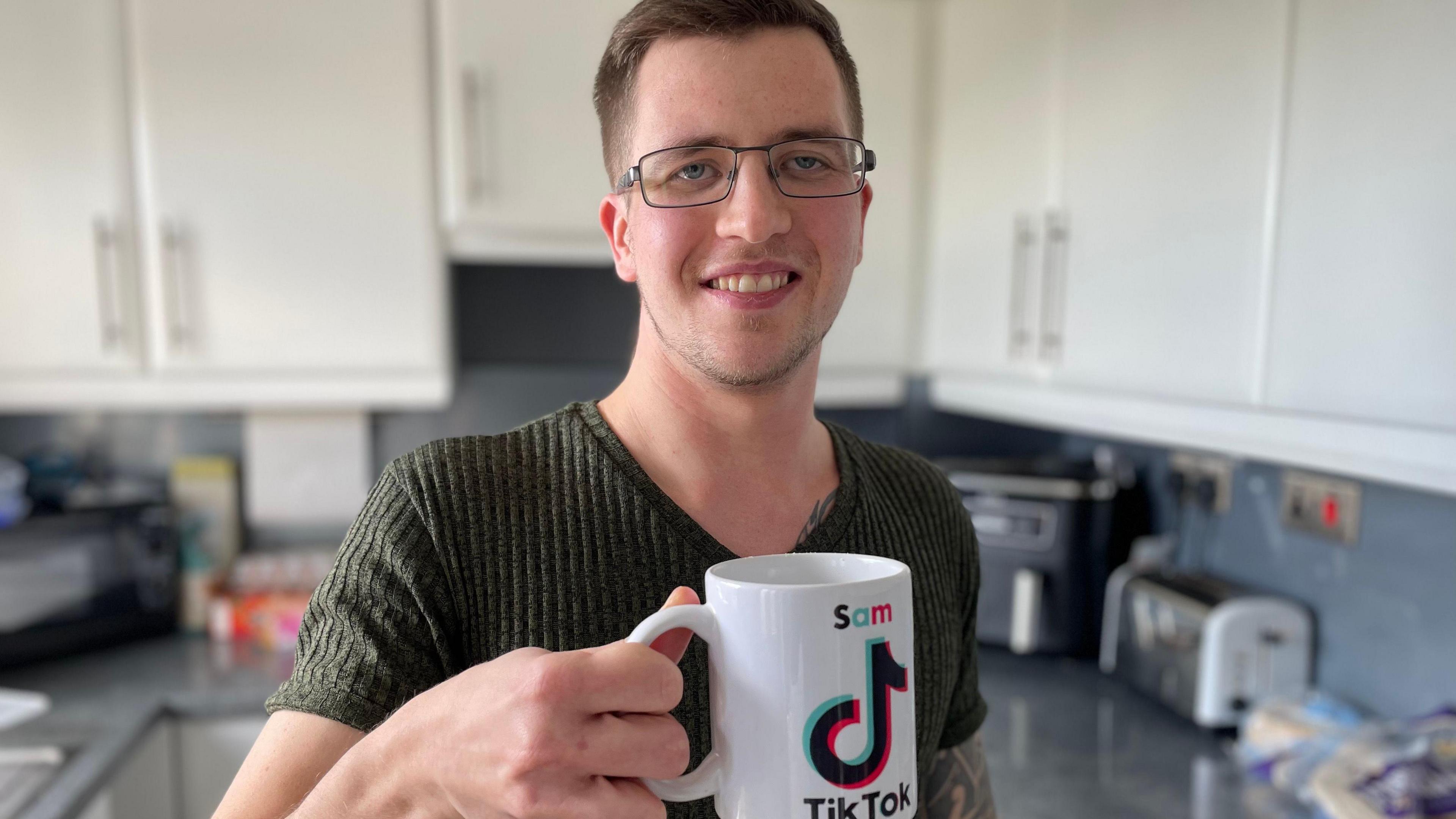 Sam Bagshaw in his kitchen, a young man with short, brown hair and glasses. He is holding a TikTok-branded mug with his name on it.