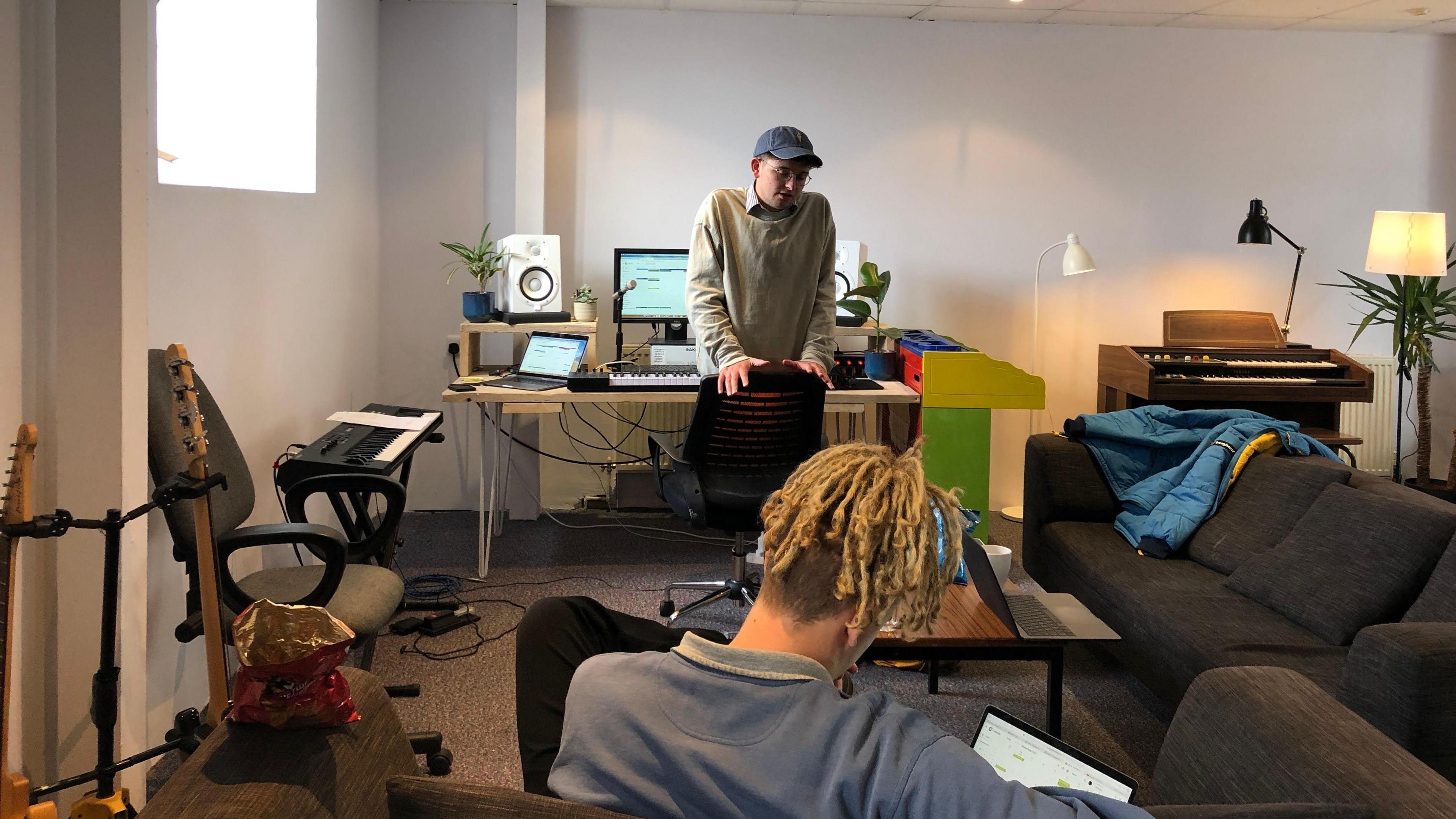 George Moir is in the studio at Burton Chapel Records. He is leaning on a chair, which is near a desk covered in computers and recording equipment. There are two dark grey sofas, where a man sits with his back to the camera, but he has blond dreadlocks tied up in a ponytail and wears a blue sweatshirt. There are guitars on stands to the left of the image and a piano to the far right, next to a lamp and plant.