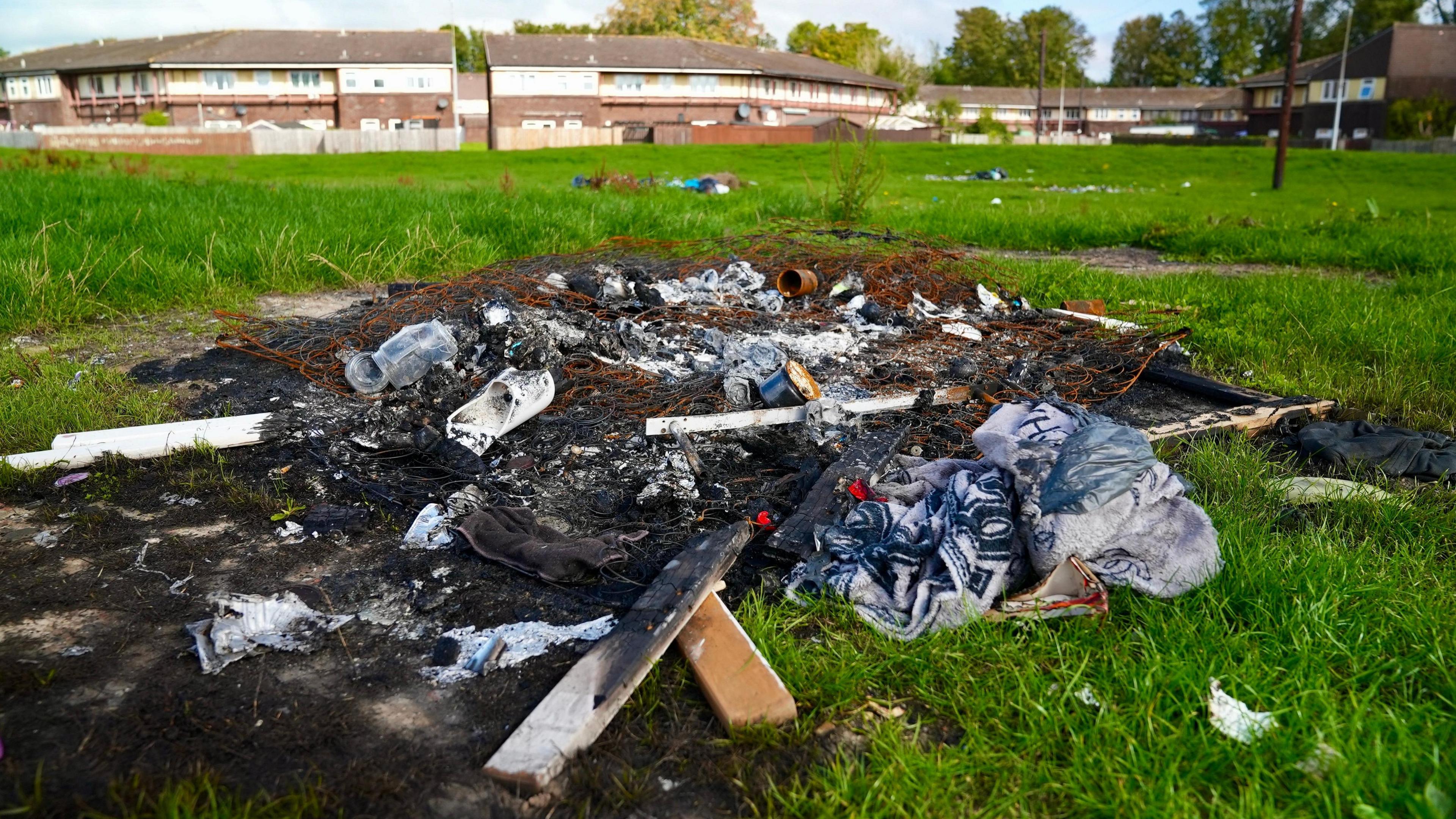 A pile of burnt rubbish in the foreground, including what's left of a burnt mattress, you can see more rubbish and then homes in the distance.