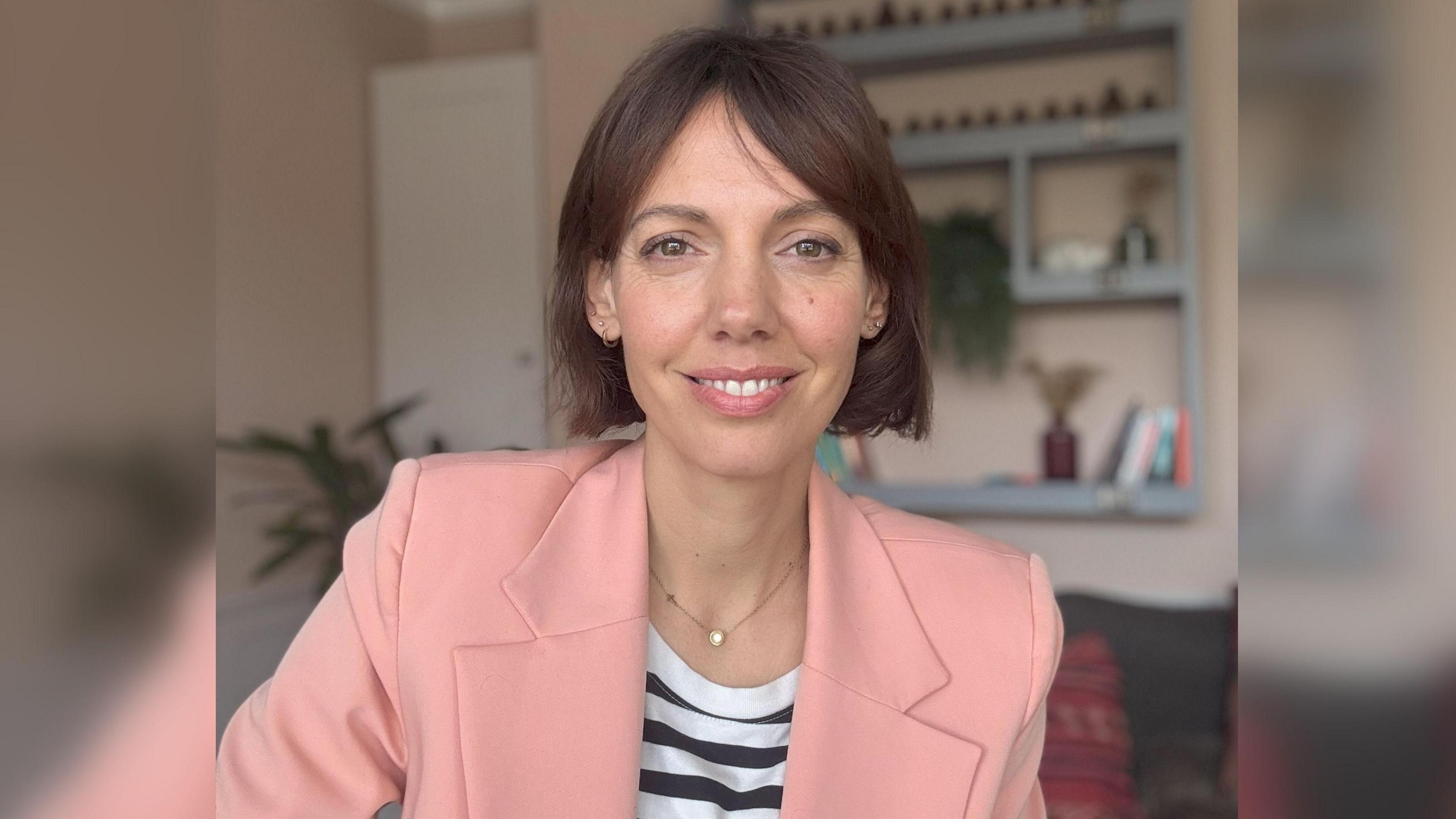 Dani Billington is the founder of the charity Menopause and Cancer. She has brown hair and is wearing a light pink blazer and a black and white striped top. She is sitting in her living room. 
