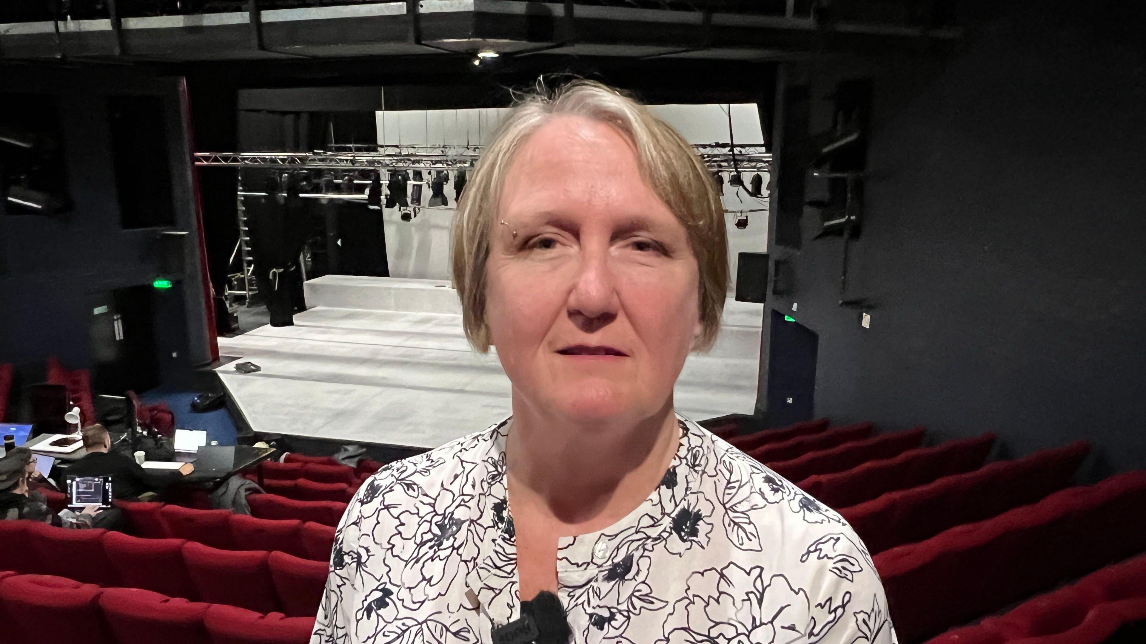 A woman is standing looking into the camera with a neutral expression. She's wearing a white blouse with the outline of flowers on. In the background are red seats facing towards a white stage.