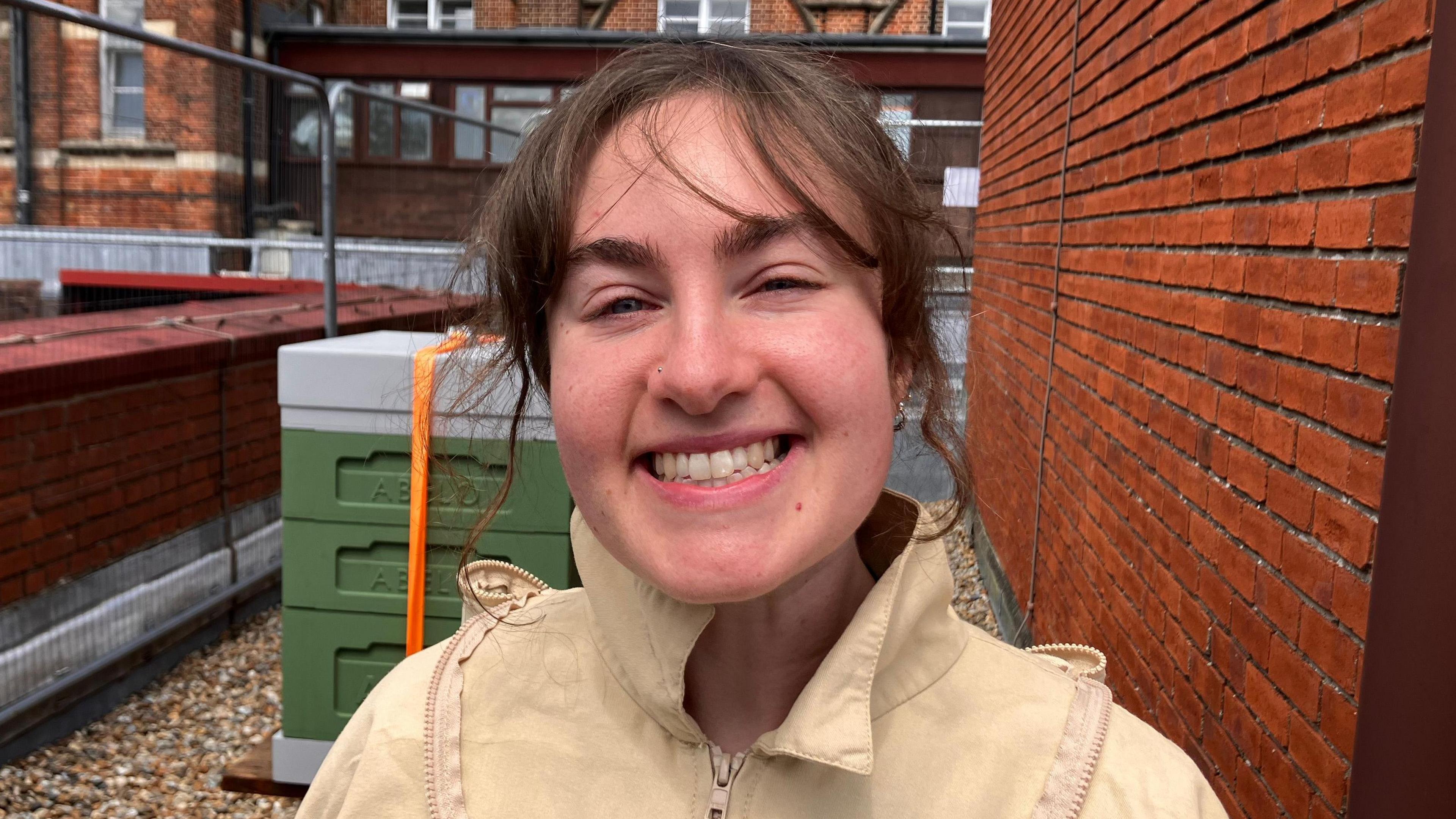 Megan wears a beekeeping suit, and has brown hair tied up. She looks excited, the hives are behind her.