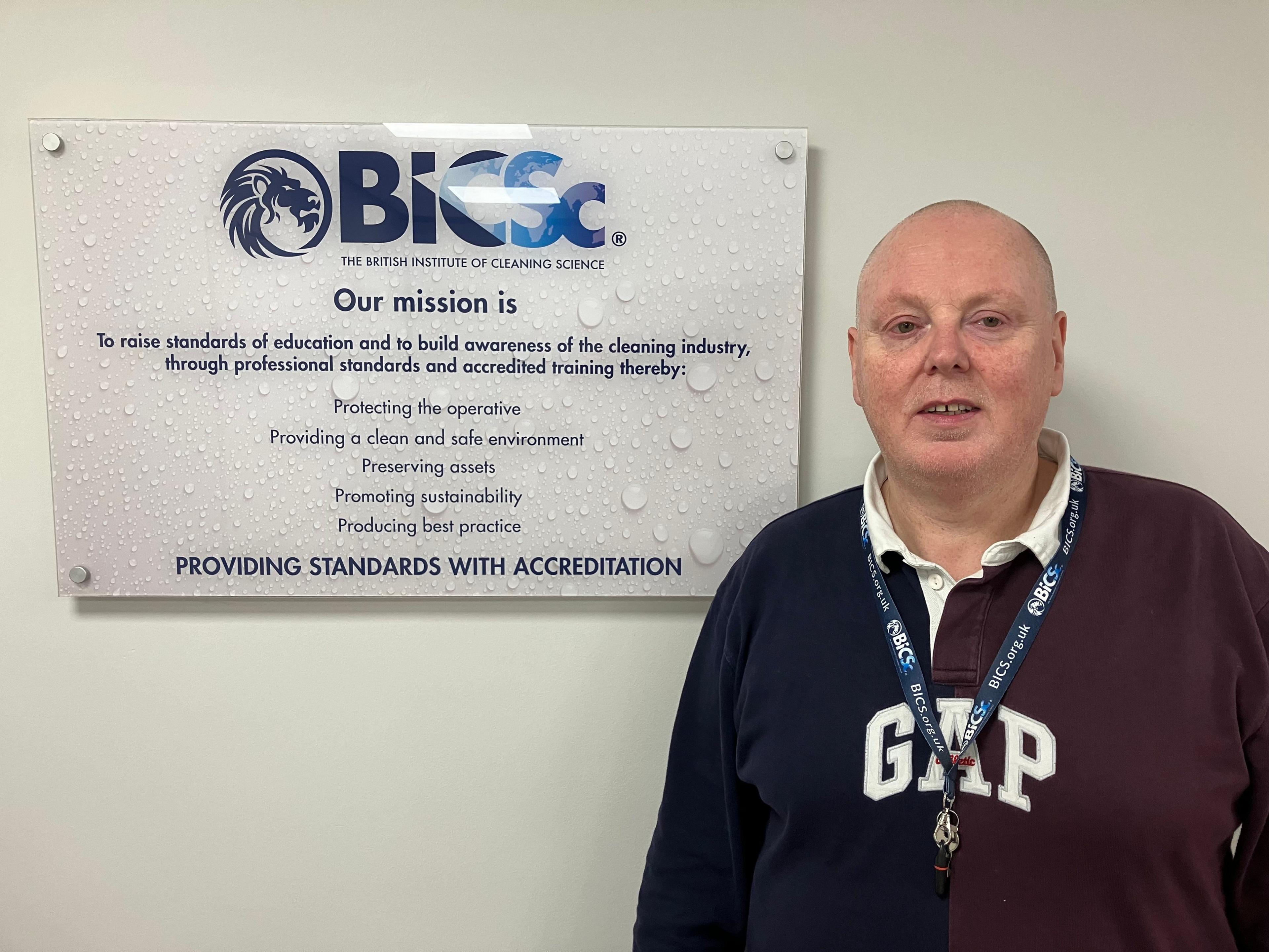 A man in a navy long sleeve polo shirt stands in front of the British Institute of Cleaning Science logo