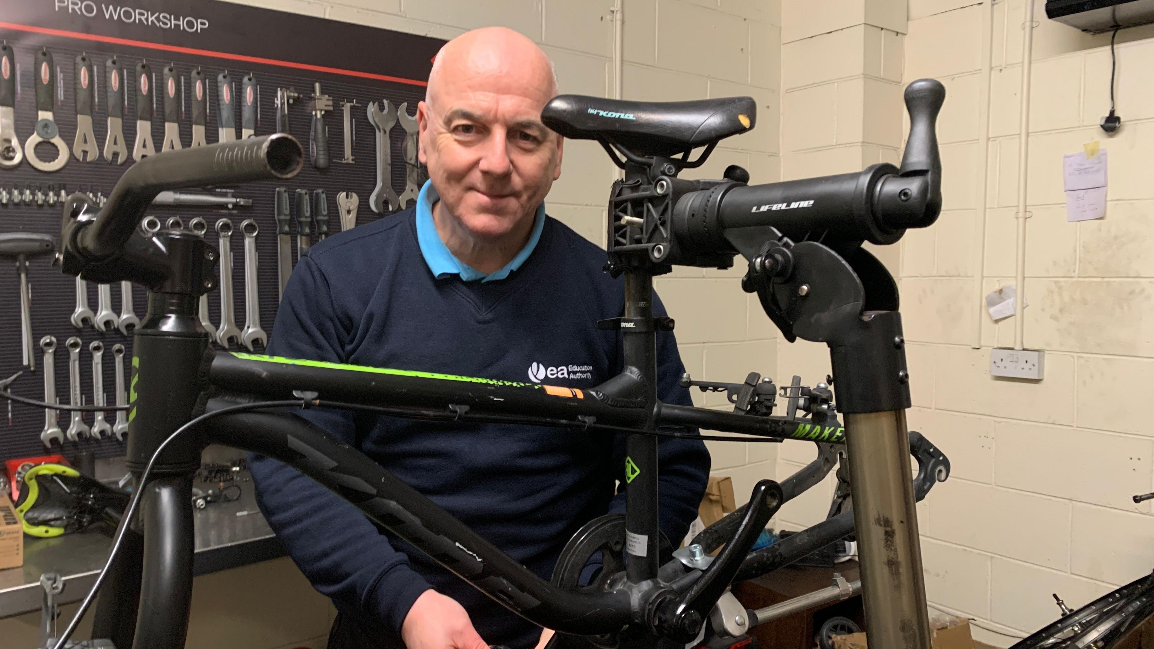 A bald man looks into the camera smiling, he's wearing a navy blue jumper with a bright blue polo neck top below it. He is standing behind a black bike. Behind him a number of silver-coloured tools are on the wall. 