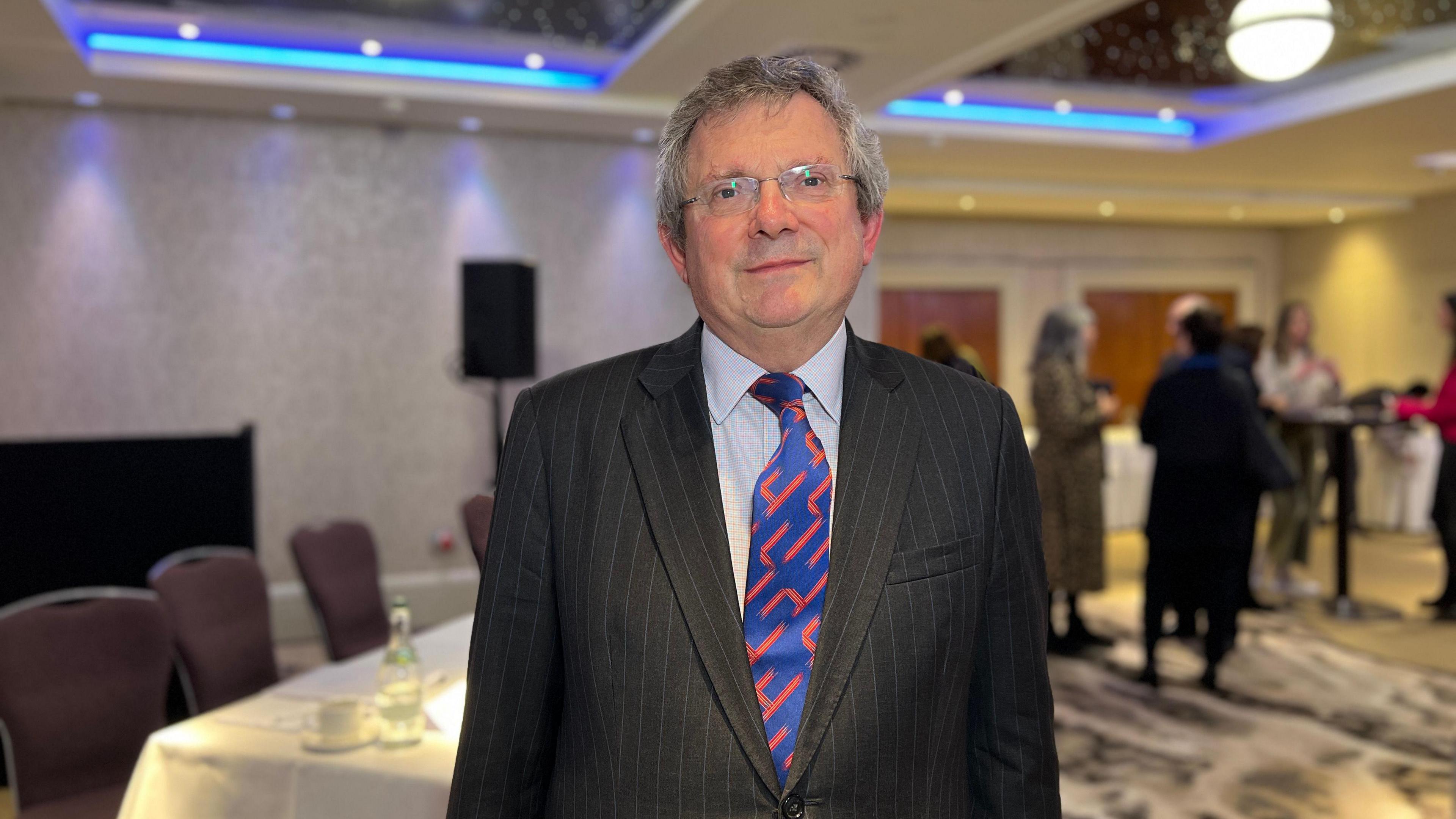 Charity commissioner John Mills looks at the camera wearing a suit as people stand behind him talking to each other after the conference finished