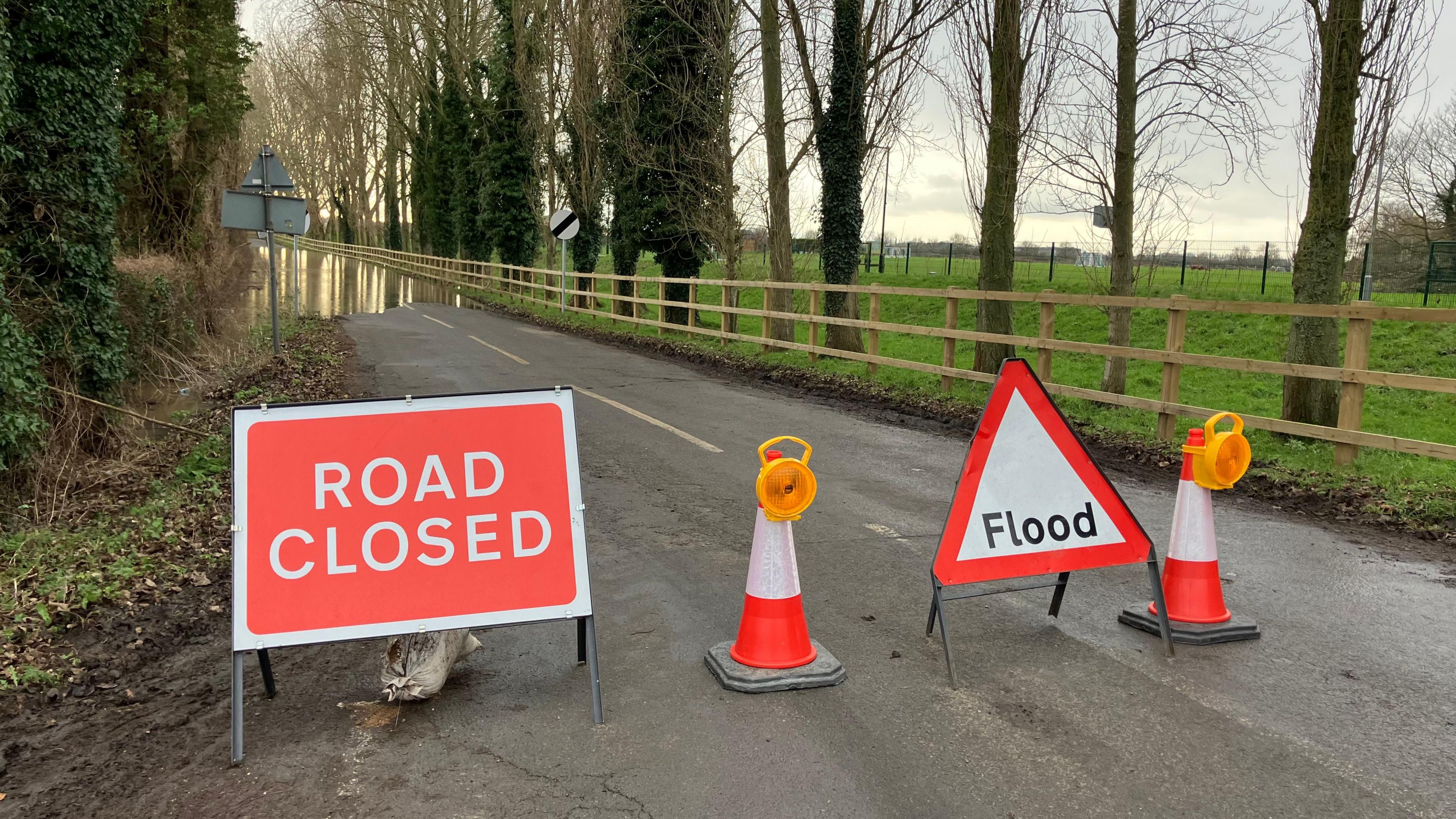 A rural road, with cones and two signs - one reading Road Closed and the other Flood - with flood water in the distance