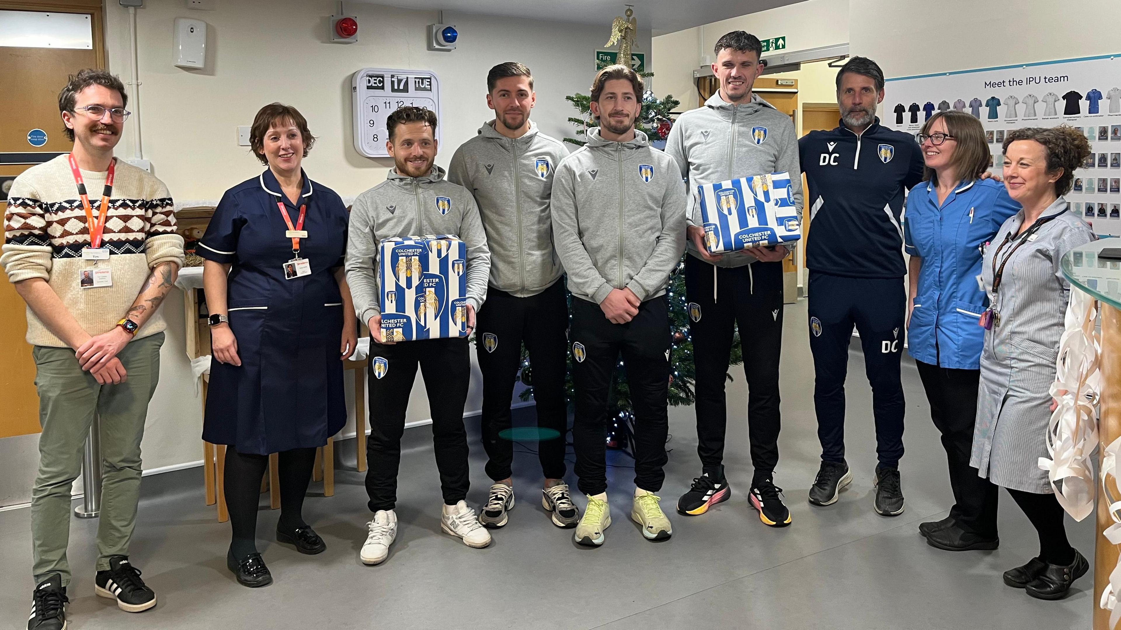 Footballers posing in front of a Christmas tree with the staff. Two of the footballers are holding wrapped Christmas gifts. 
Everyone is smiling at the camera