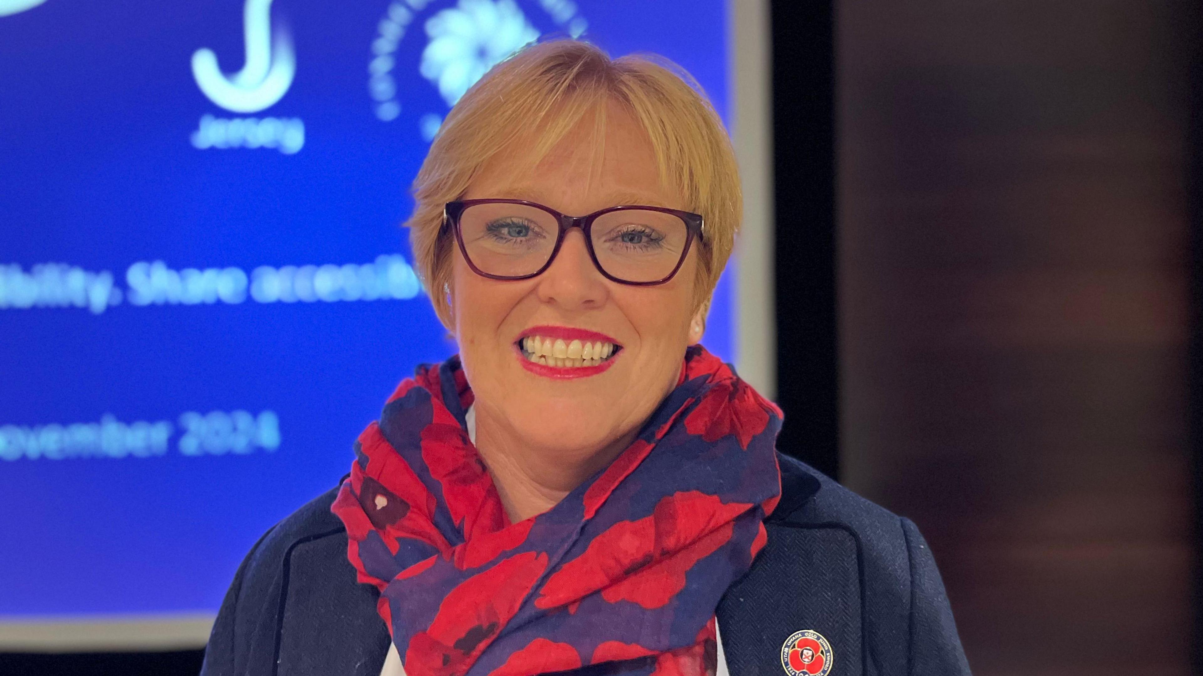 Cirsty de Gruchy Moseley is a woman with short blonde hair. She is wearing a red and purple scarf with poppies on and a navy blazer. She has a poppy pin on her right lapel. She is wearing red glasses and pink lipstick. She is smiling at the camera. The background is an out of focus projector, with a purple slide on it.