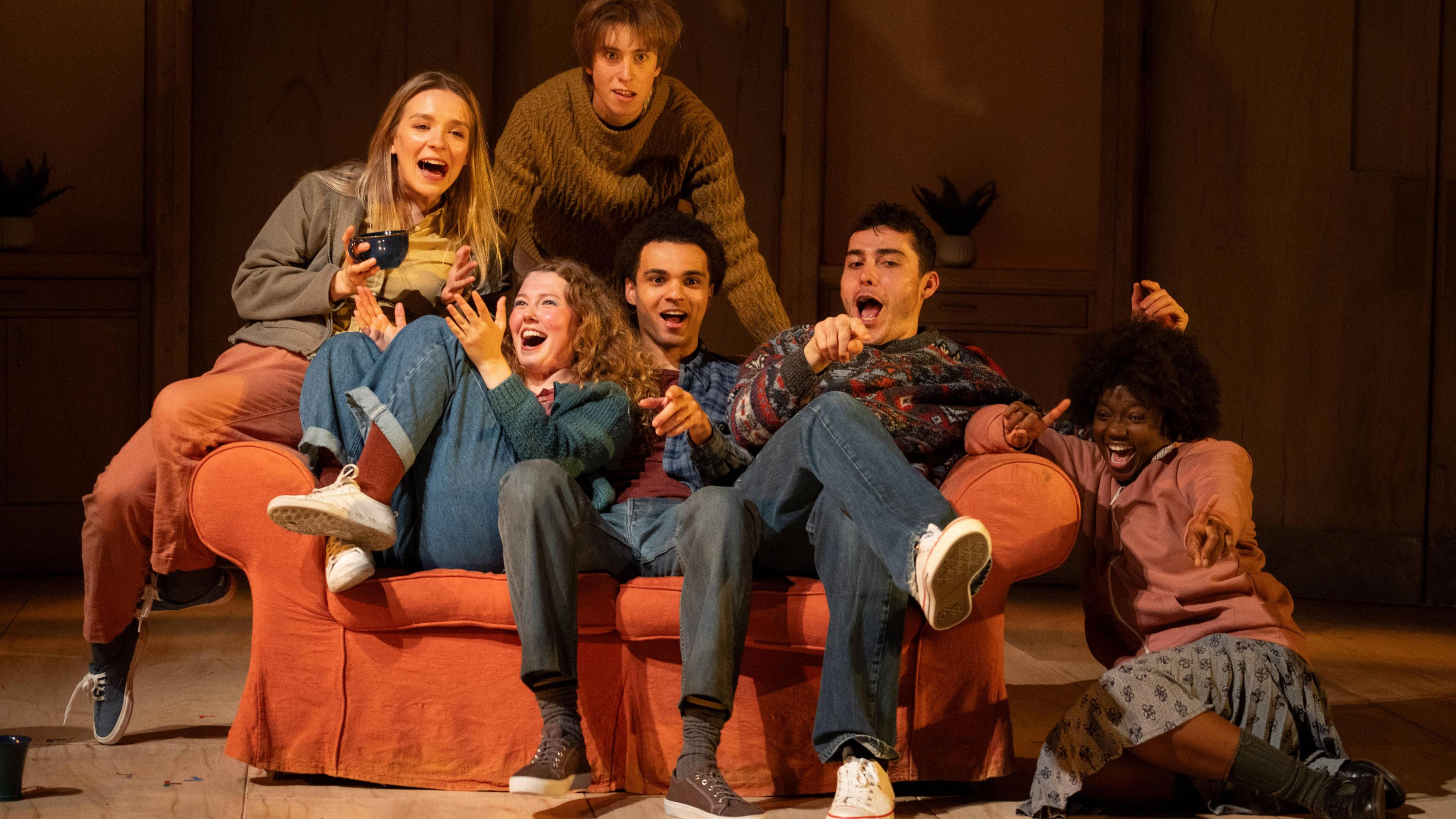 The cast of Never Let Me Go - a group of young actors. The picture is taken during a performance of the play. The actors are gathered on and around a sofa and are smiling and laughing.