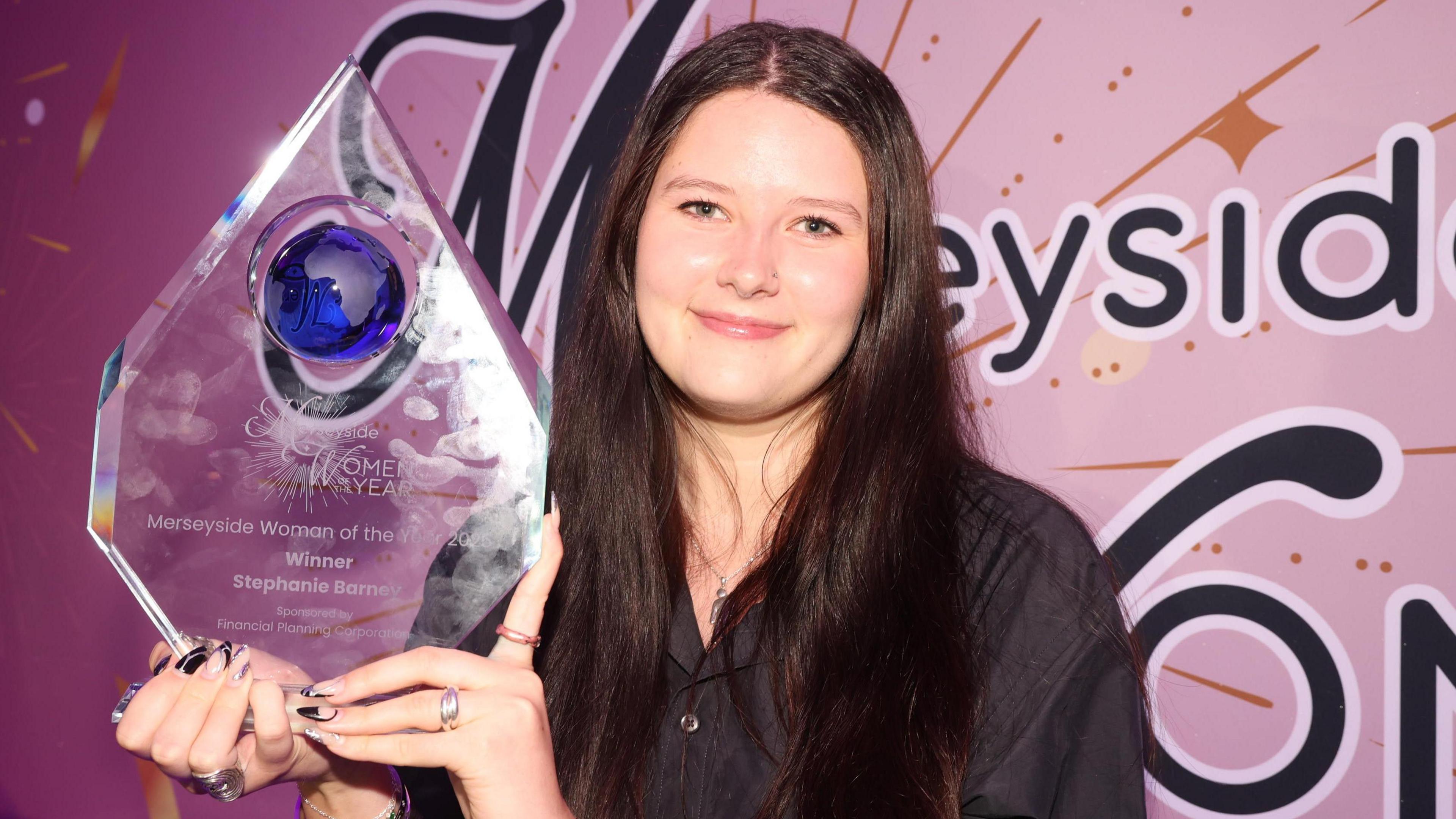 Stephanie Barney has long, dark brown hair and smiles for the cameras while holding her large trophy for winning the Merseyside Woman of the Year award.