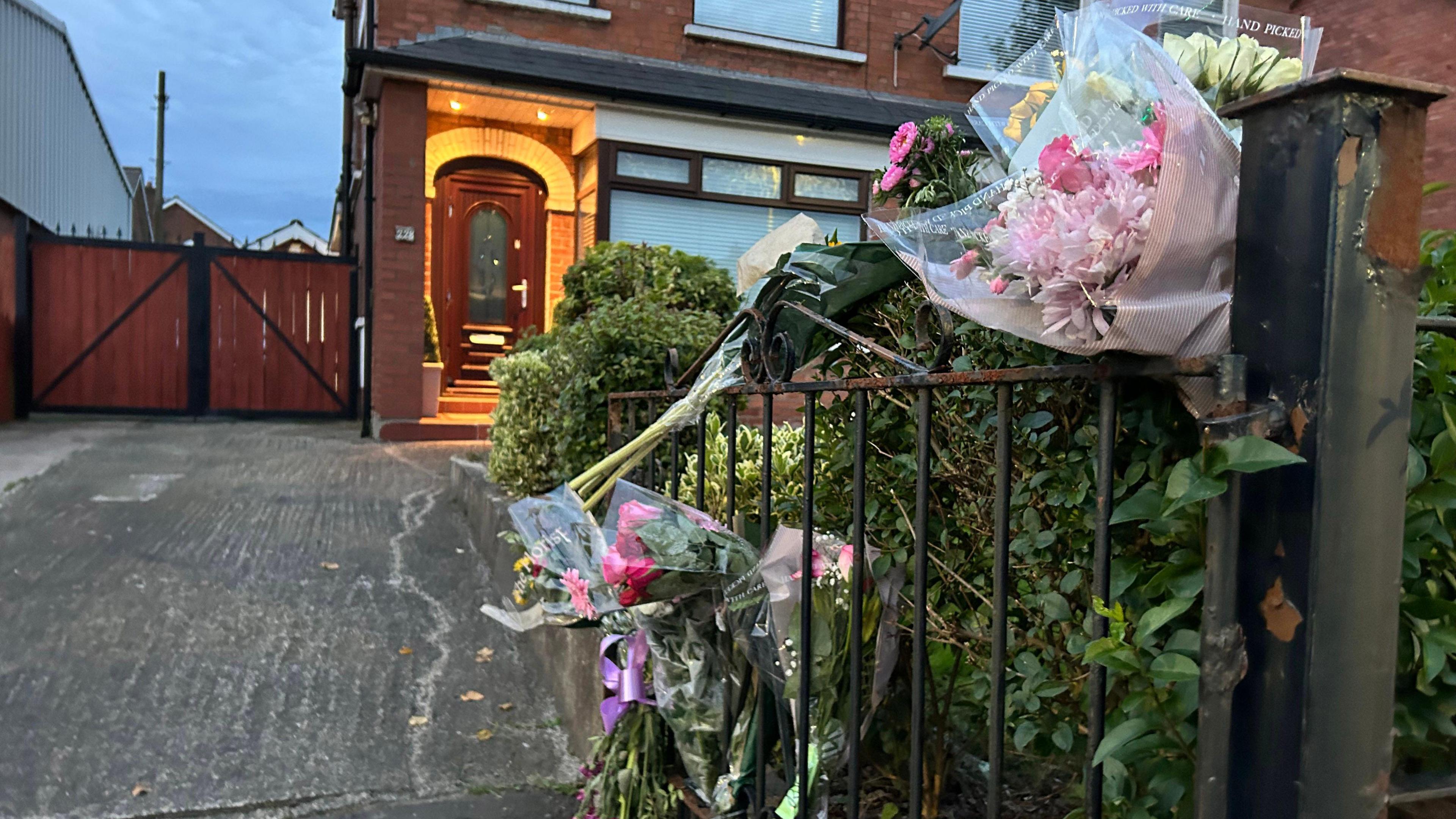 An open gate leading to a front door on a red brick house. There are bunches of flowers attached to the gate. A window in the background has the blind drawn down.