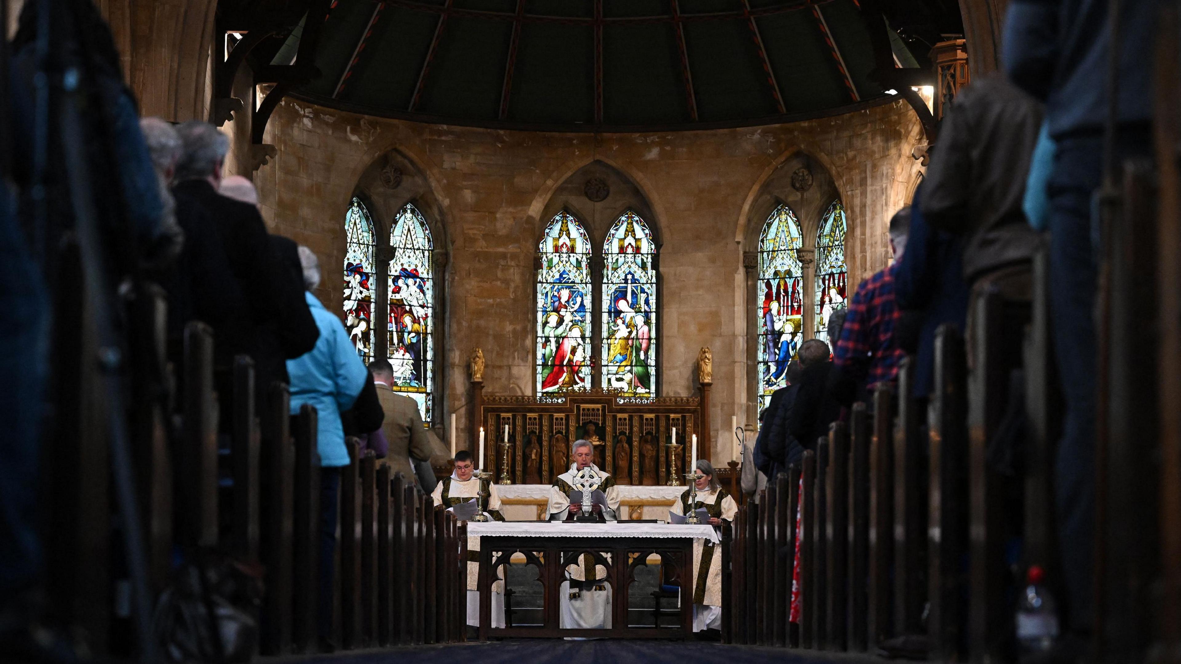 A ceremony takes place at the Holy Trinity Church in Llandudno, north Wales. There are rows of people sitting in church pews seen the left and right with the photo being taken at ground level from the central corridor as you enter the church. Archbishop of Wales Andrew John is stood behind a cross in the image, in front of the stained glass windows and walls of the church.