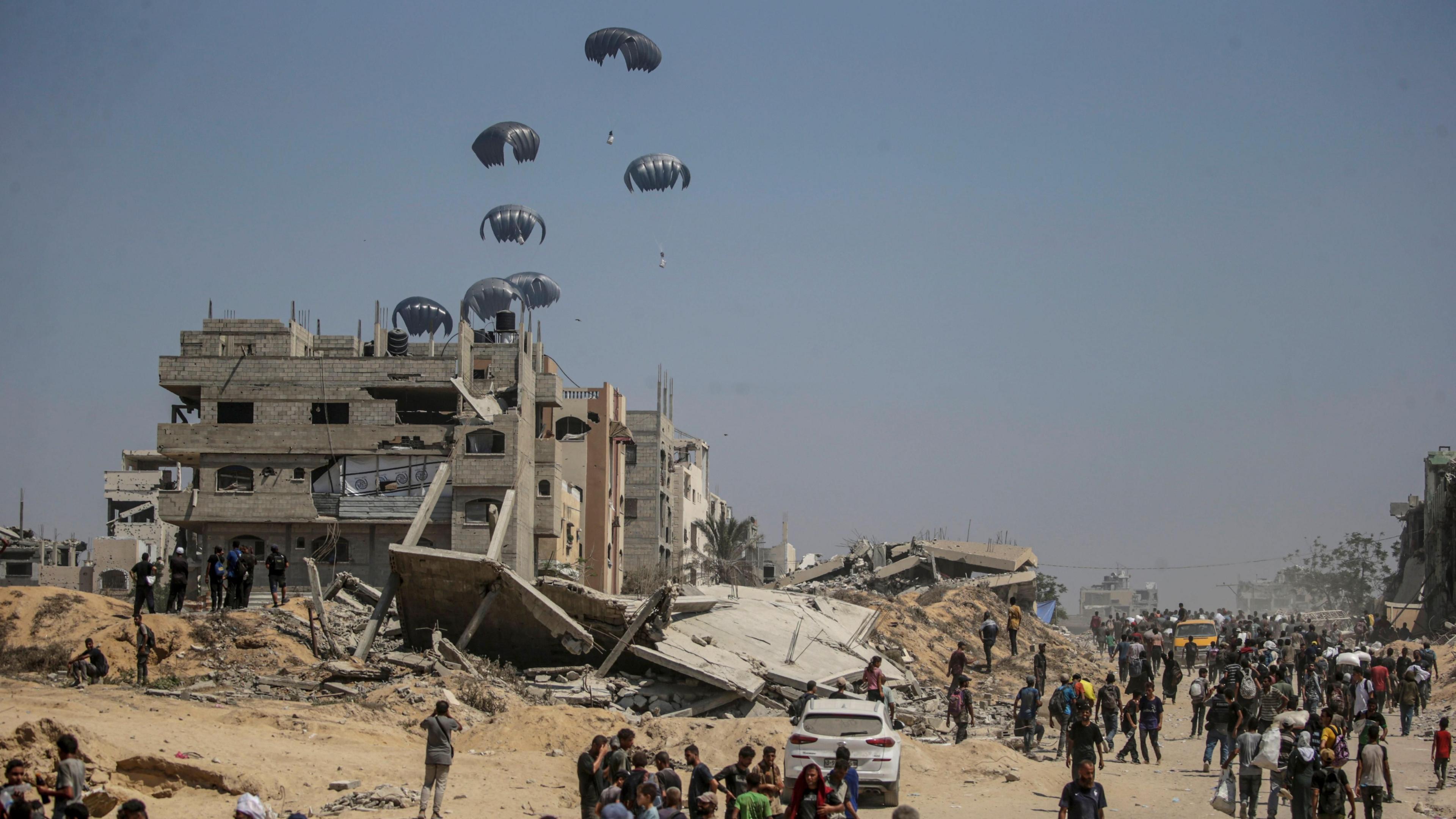 Humanitarian aid airdropped by the Royal Jordanian Army lands in the northern Gaza Strip. Silvery parachutes can be seen descending near several damaged buildings.