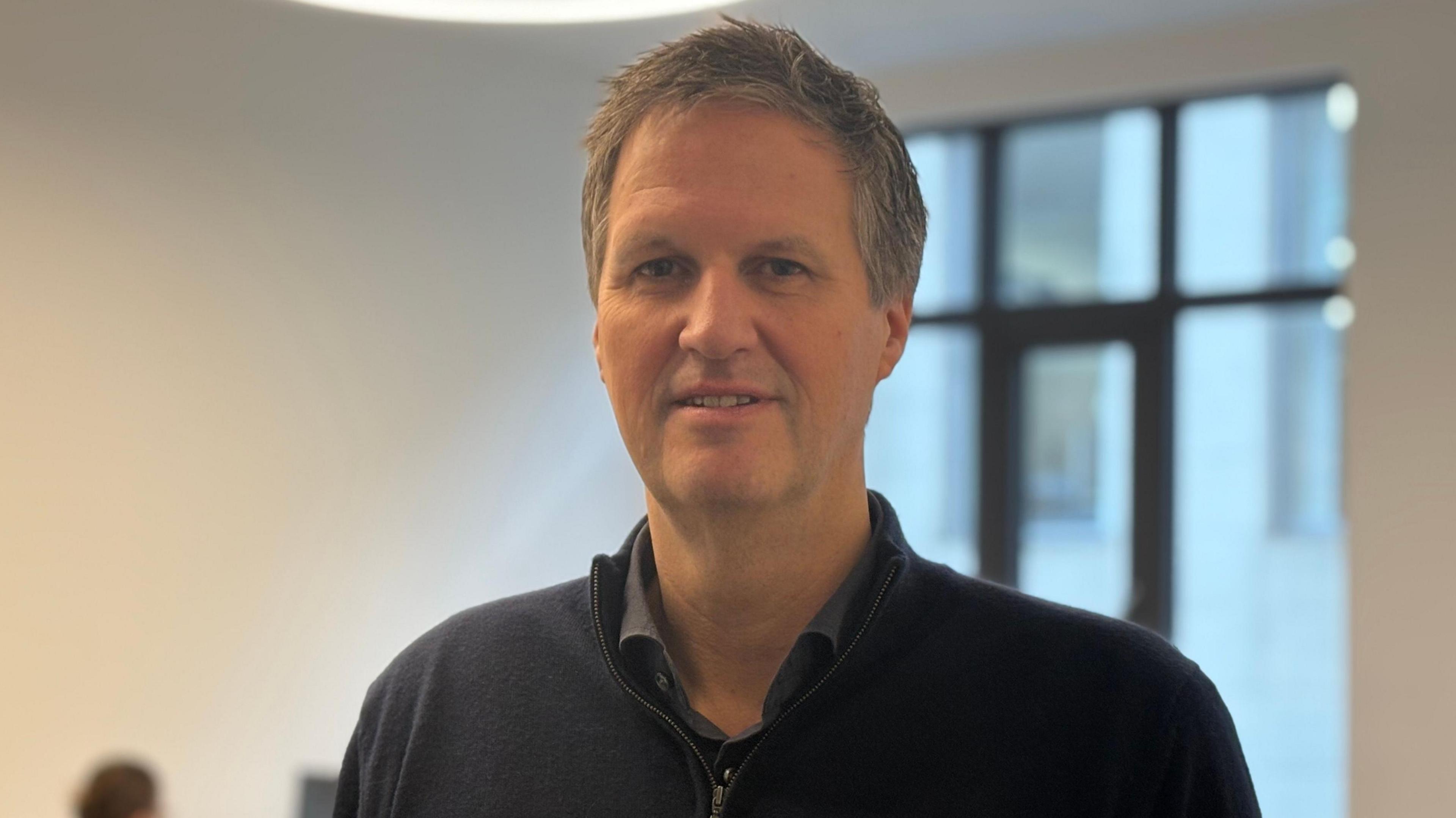 Greg Watson, Napier NI CEO, is standing in a white room with a large window behind him. He has short grey hair and is wearing a dark coloured zip jumper.