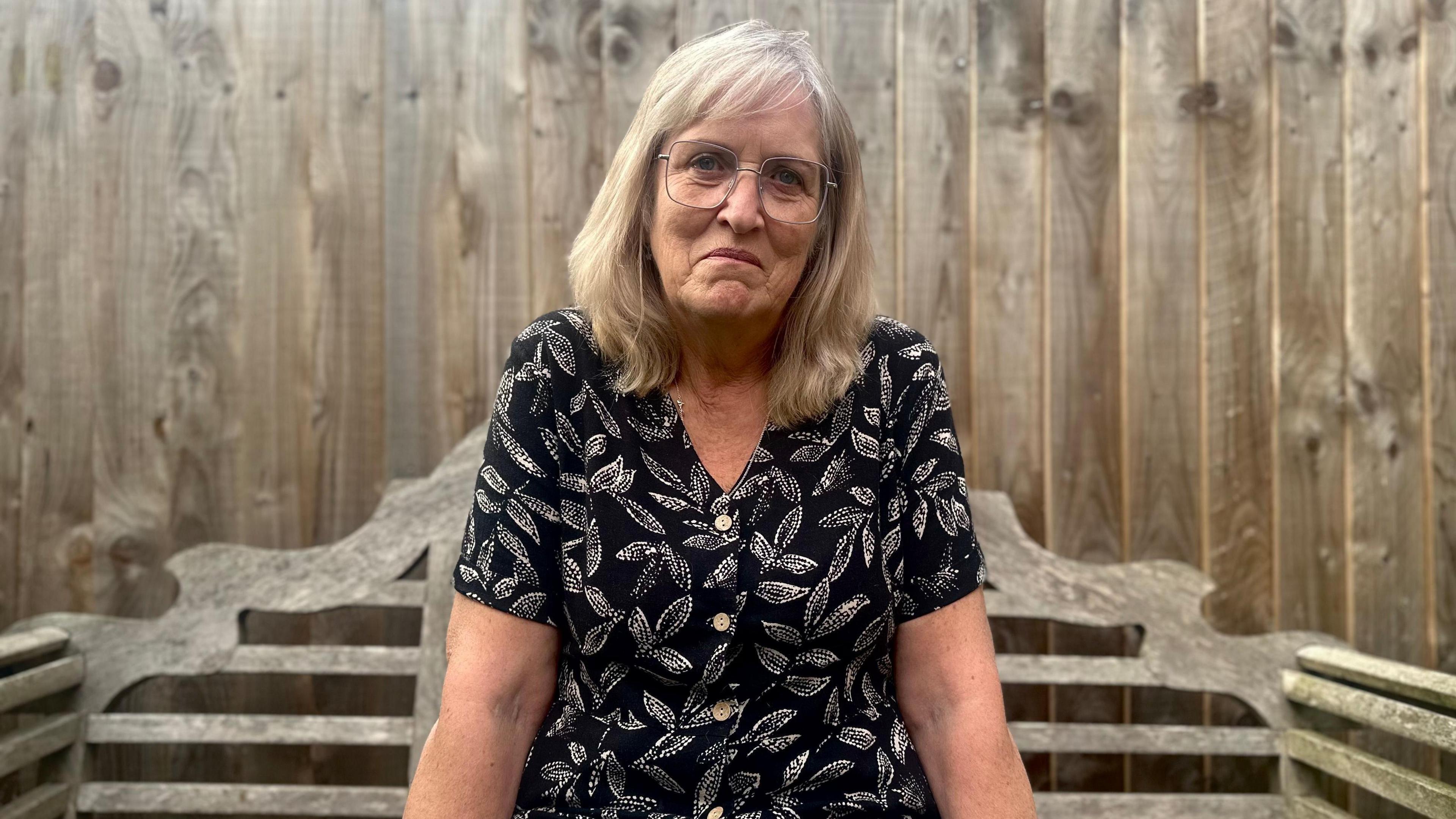 Nicki Duffield sitting on a wooden bench looking at the camera. Behind her is a wooden fence. She is wearing a black and white leaf-pattern dress. She has blonde hair and glasses.