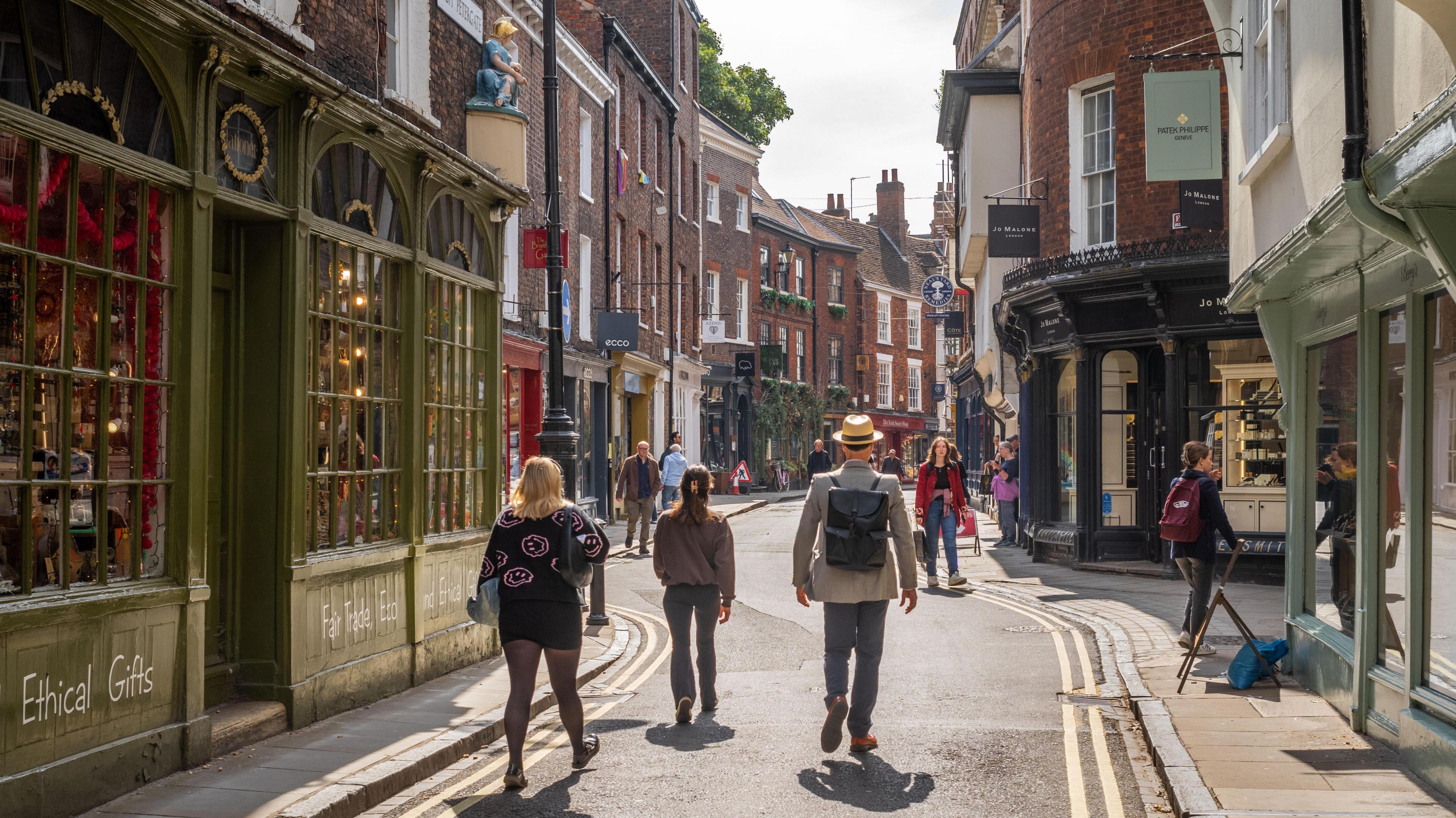 People walking down a narrow shopping street in York on a sunny day. The road has double yellow lines on either side.