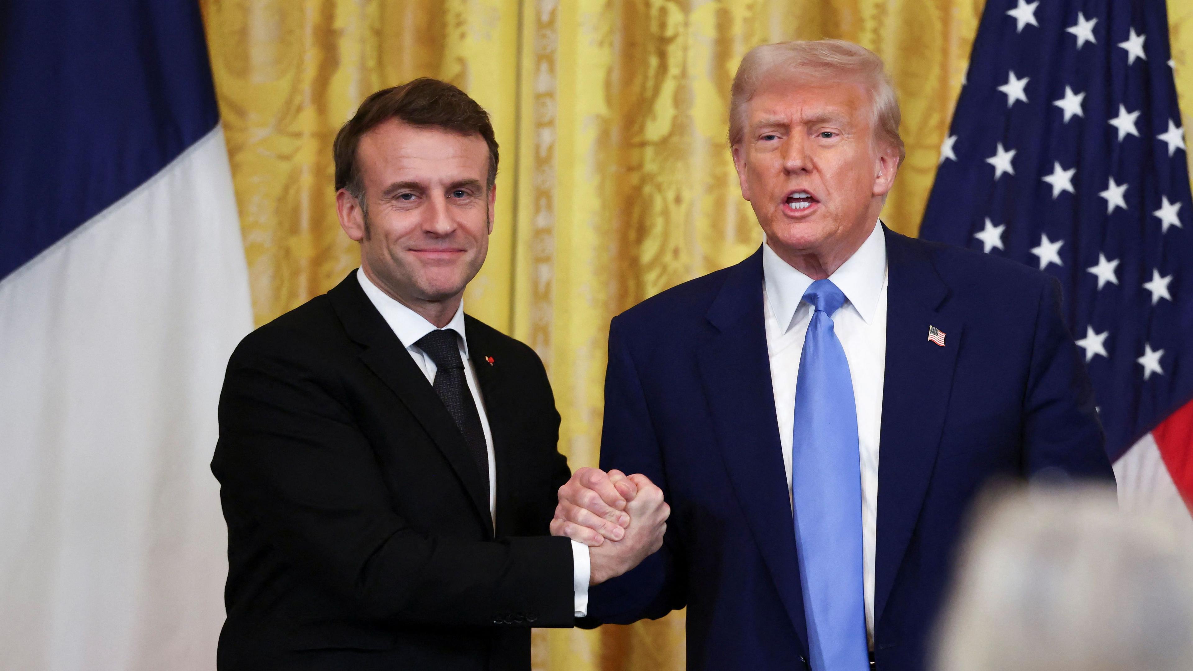 French President Emmanuel Macron and US President Donald Trump with hands clenched in a handshake in front of a French and US flag. 