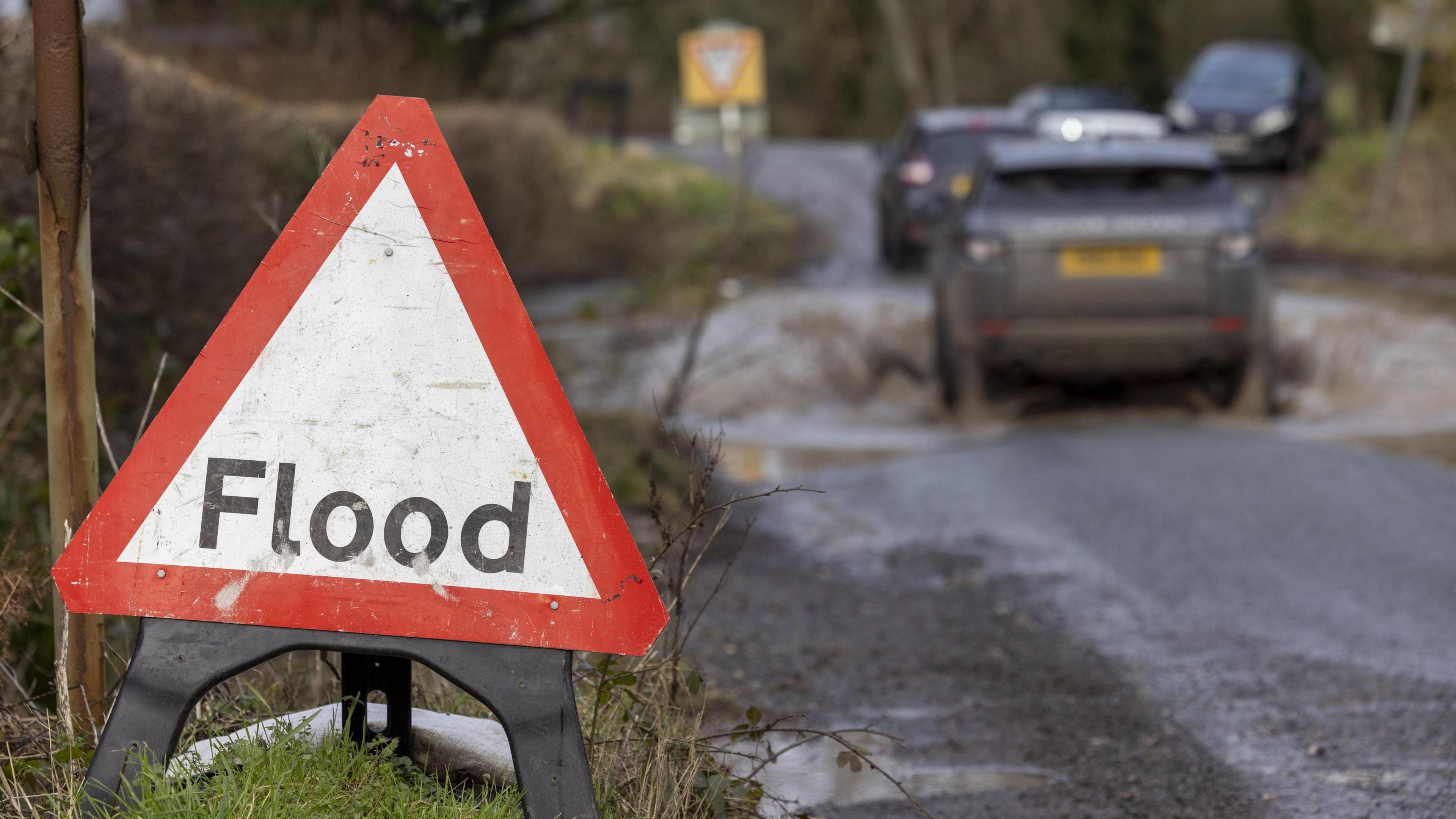 A red and white flood sign next to a road. A car can be seen driving through water. 