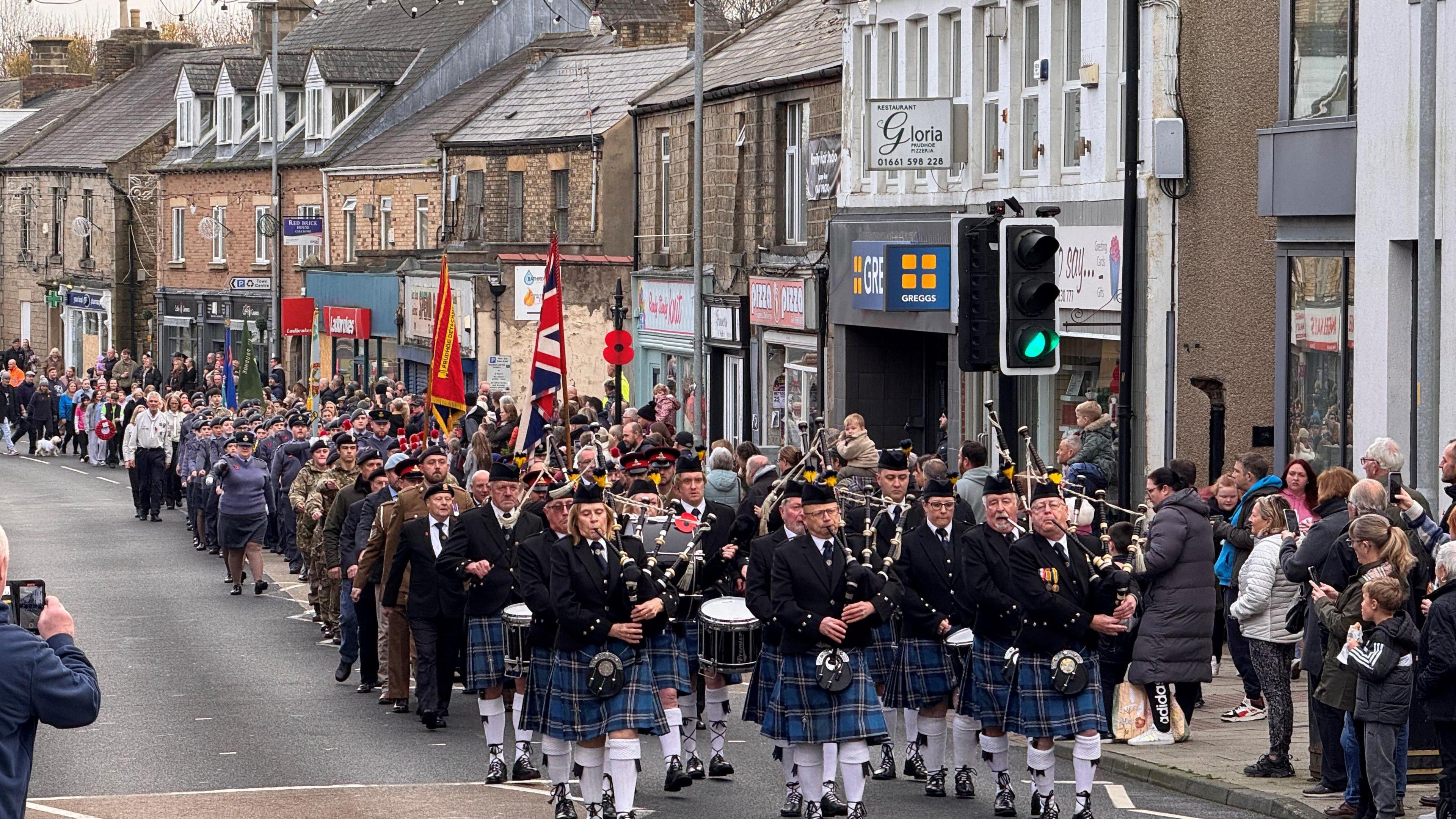 People in uniforms, including kilts, parading across the town while crowds watch from the side of the street in Prudhoe.