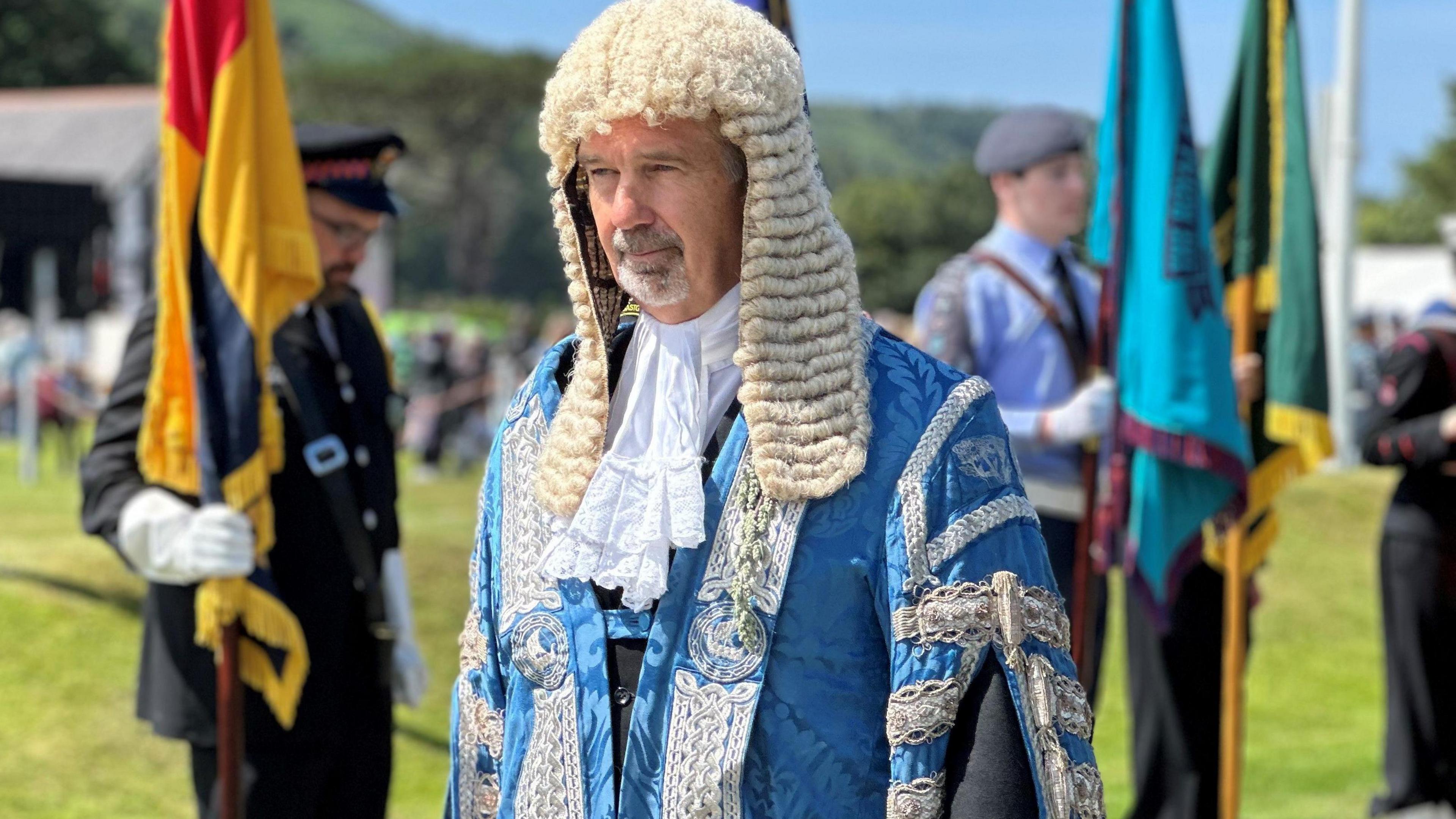 Laurence Skelly, who is wearing blue ceremonial robes and a long white wig during the procession on Tynwald Day. There are standard bearers lined up alongside the walkway behind him.