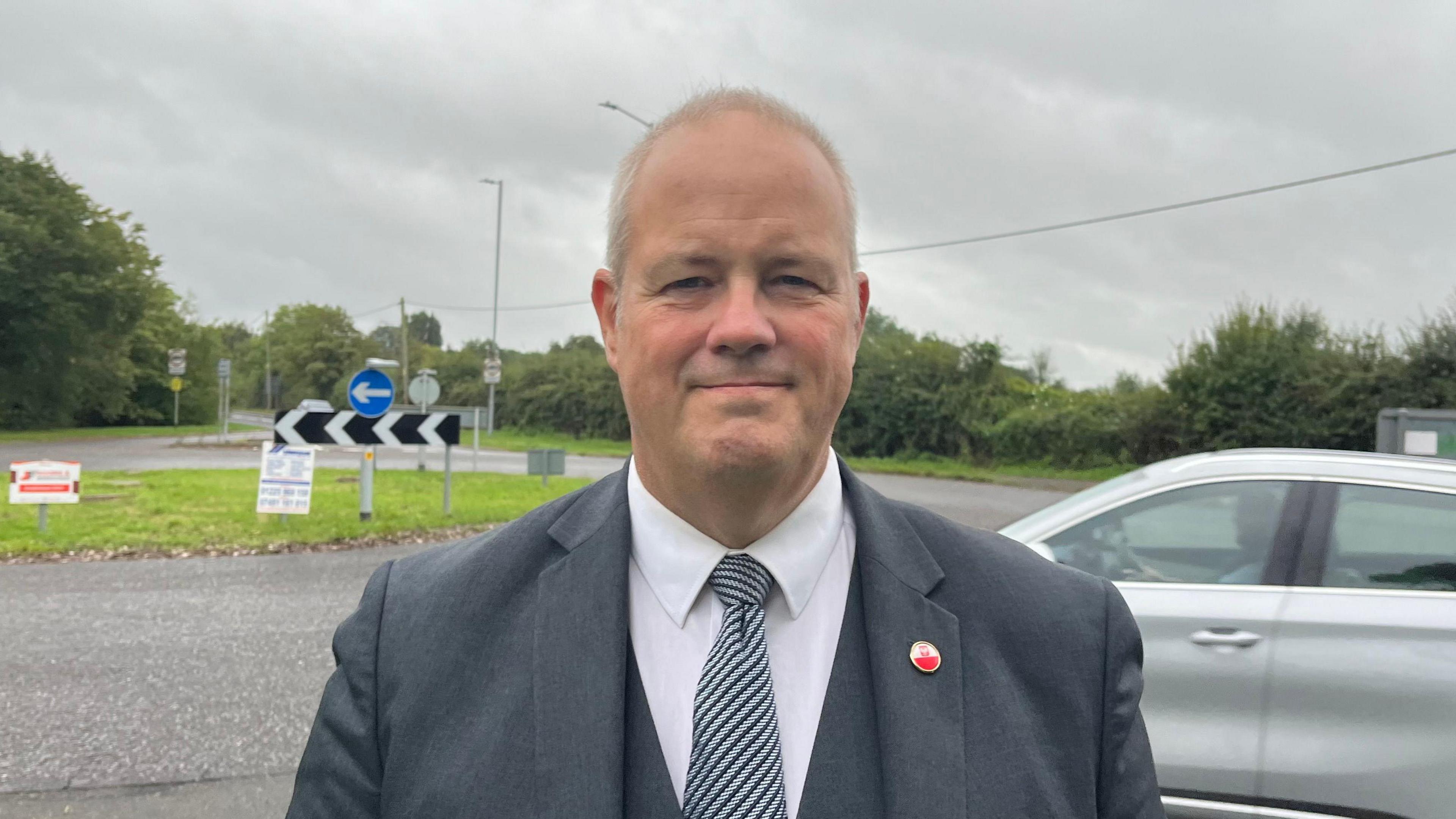 Dr Simon Selby in a suit standing in front of the roundabout looking into the camera