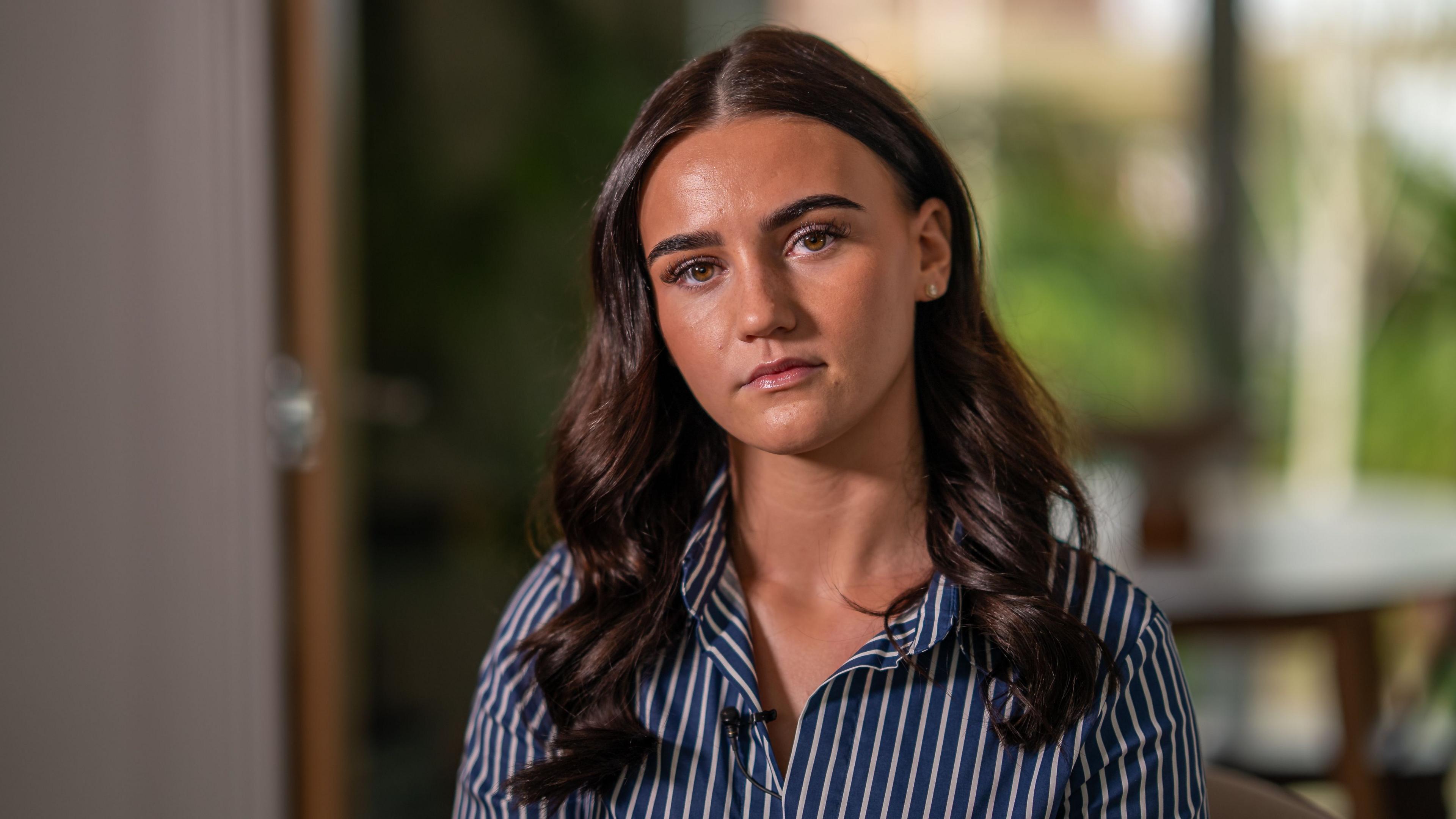 A young women with long wavy dark hair stares expressionless at the camera, wearing a blue and white striped shirt.