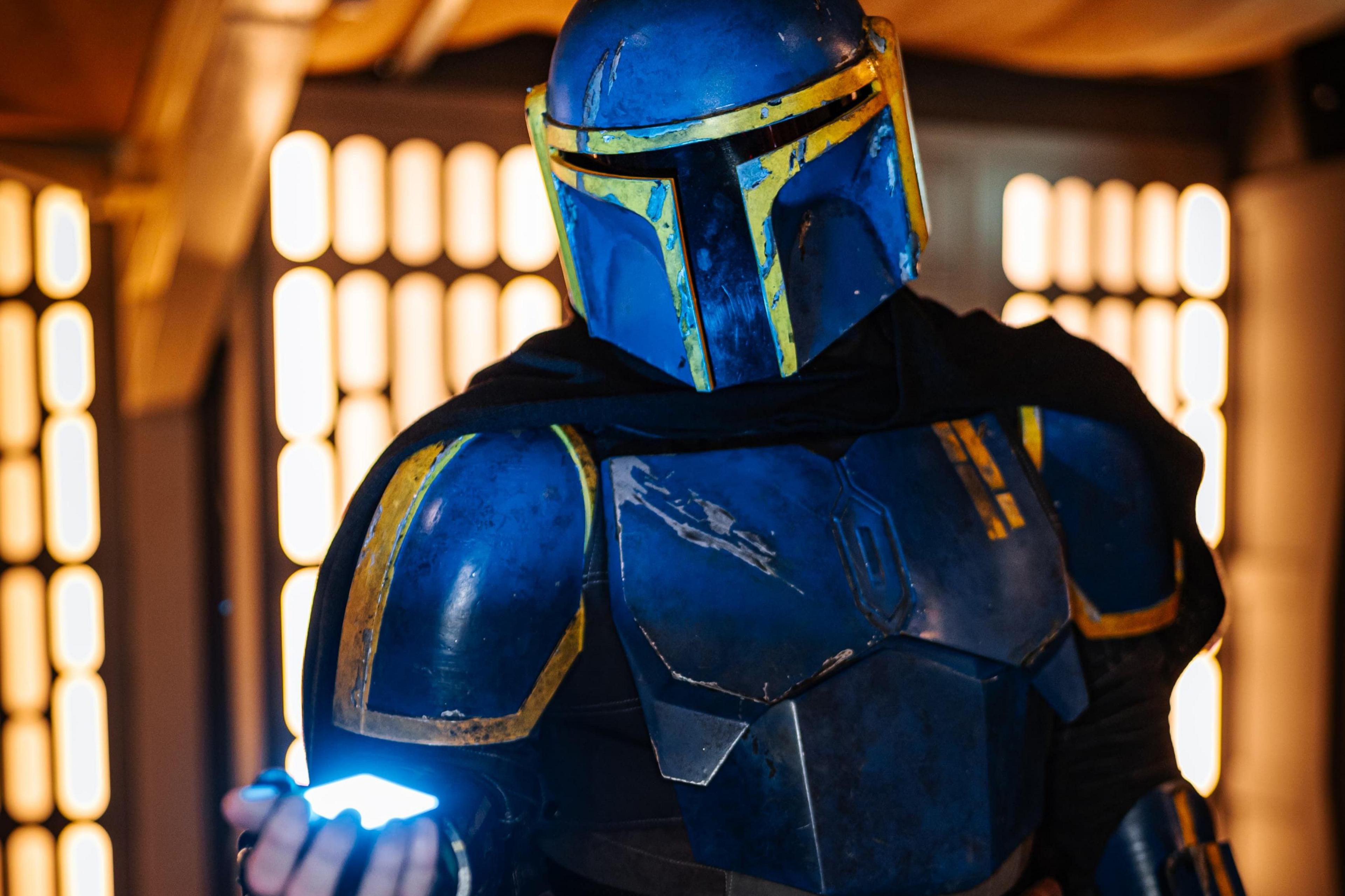 Chris stands clad in a cobalt blue distressed Mandalorian costume. He's holding a futuristic light in his right hand and is standing in front of an out-of focus tan background resembling the artistic style of many Star Wars scenes.