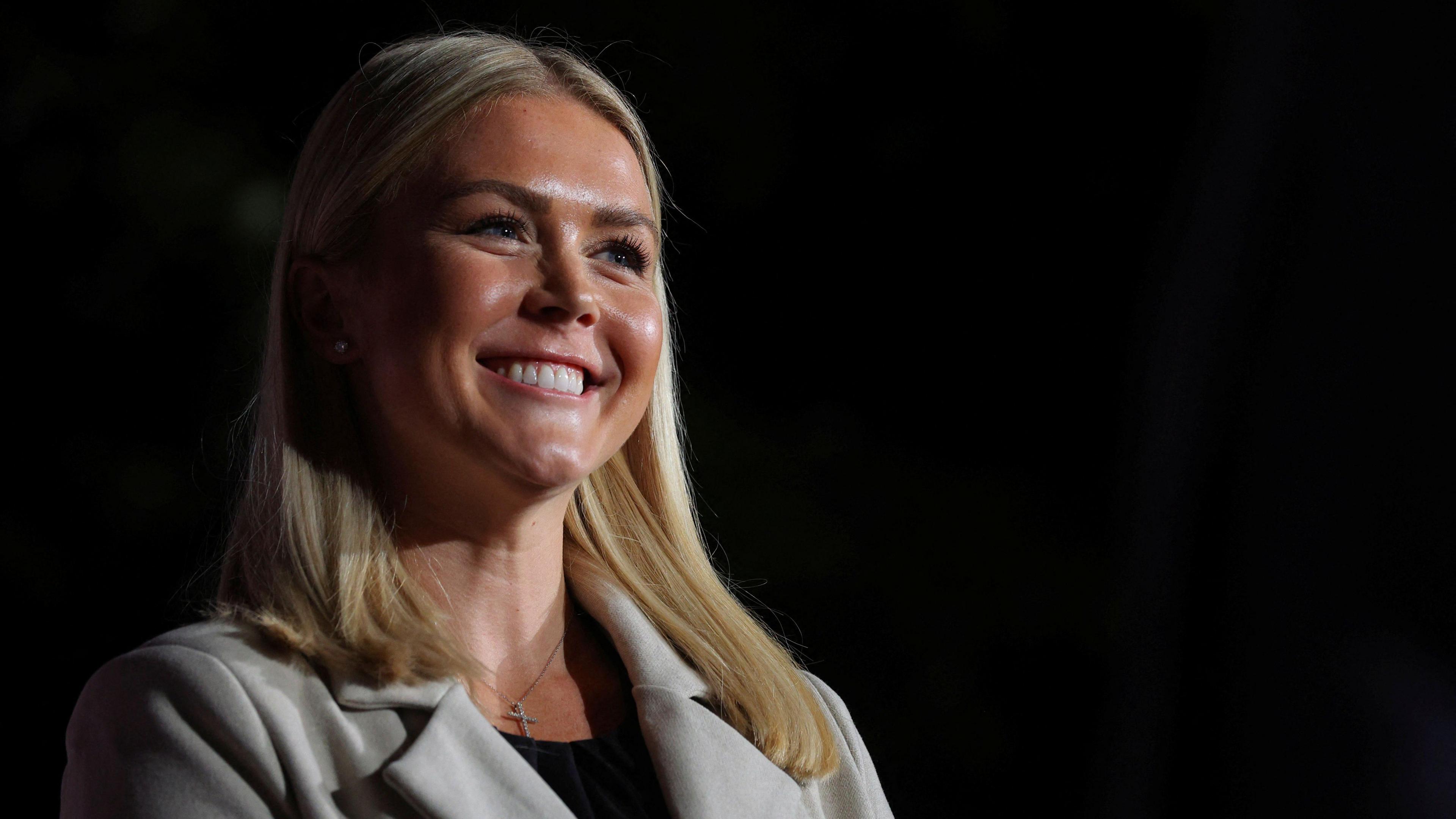 Karoline Leavitt, who has straight blonde shoulder-length hair, and wears a silver cross necklace with a cream coat, beams during a rally in 2022