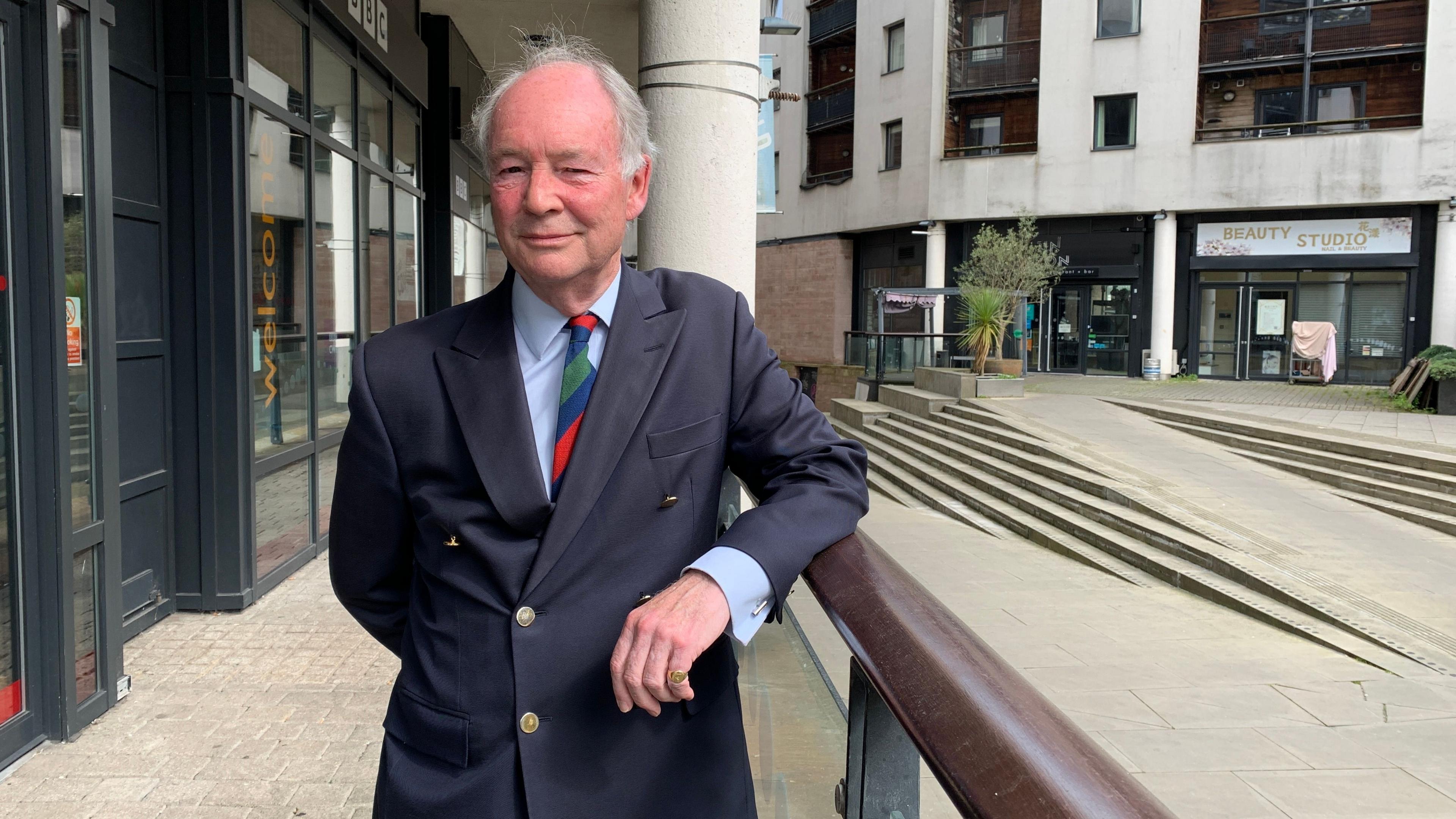 Philip Seccombe wearing a blue suit, a light blue shift and a multi-coloured tie stood in a courtyard area surrounded by tall buildings