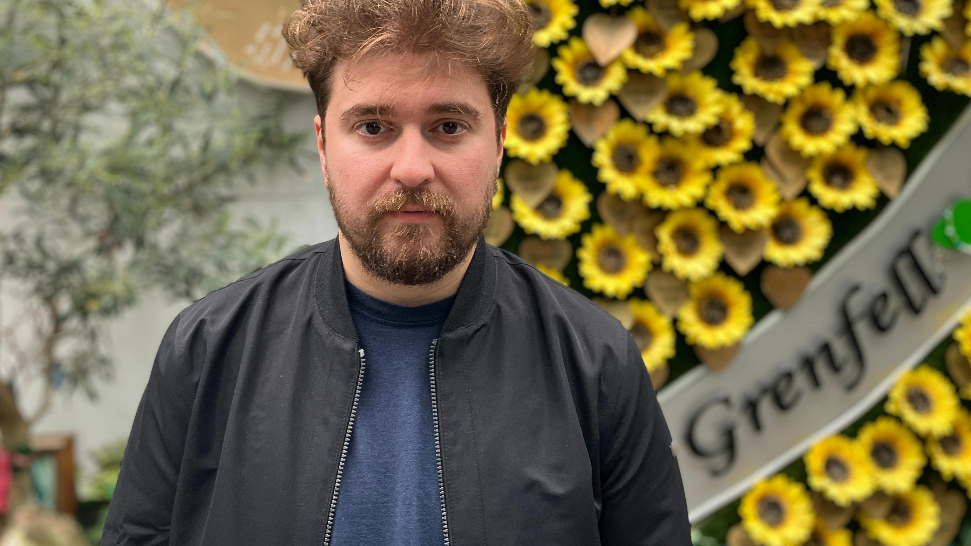 Tiago Alves wearing a black bomber jacket, blue t-shirt. He has mid-brown short hair and a full beard. He's standing in front of a Grenfell memorial sign made up of yellow sunflowers.
