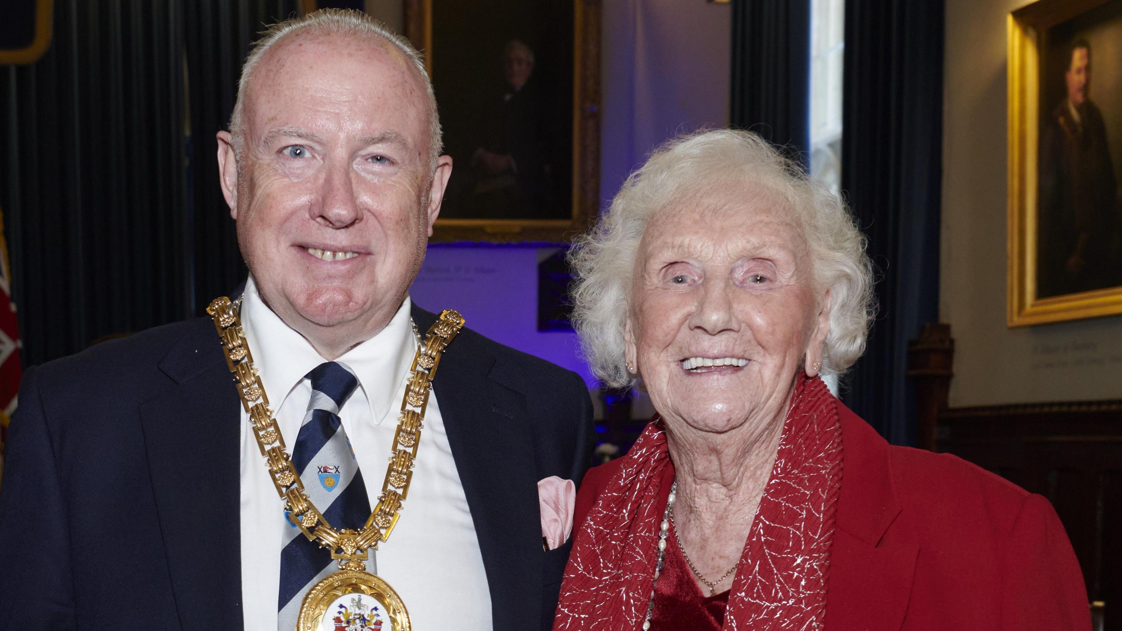 Kieron Mallon, Mayor of Banbury stood with Ena Bowley in a town hall with old portraits behind them as they smile at the camera.