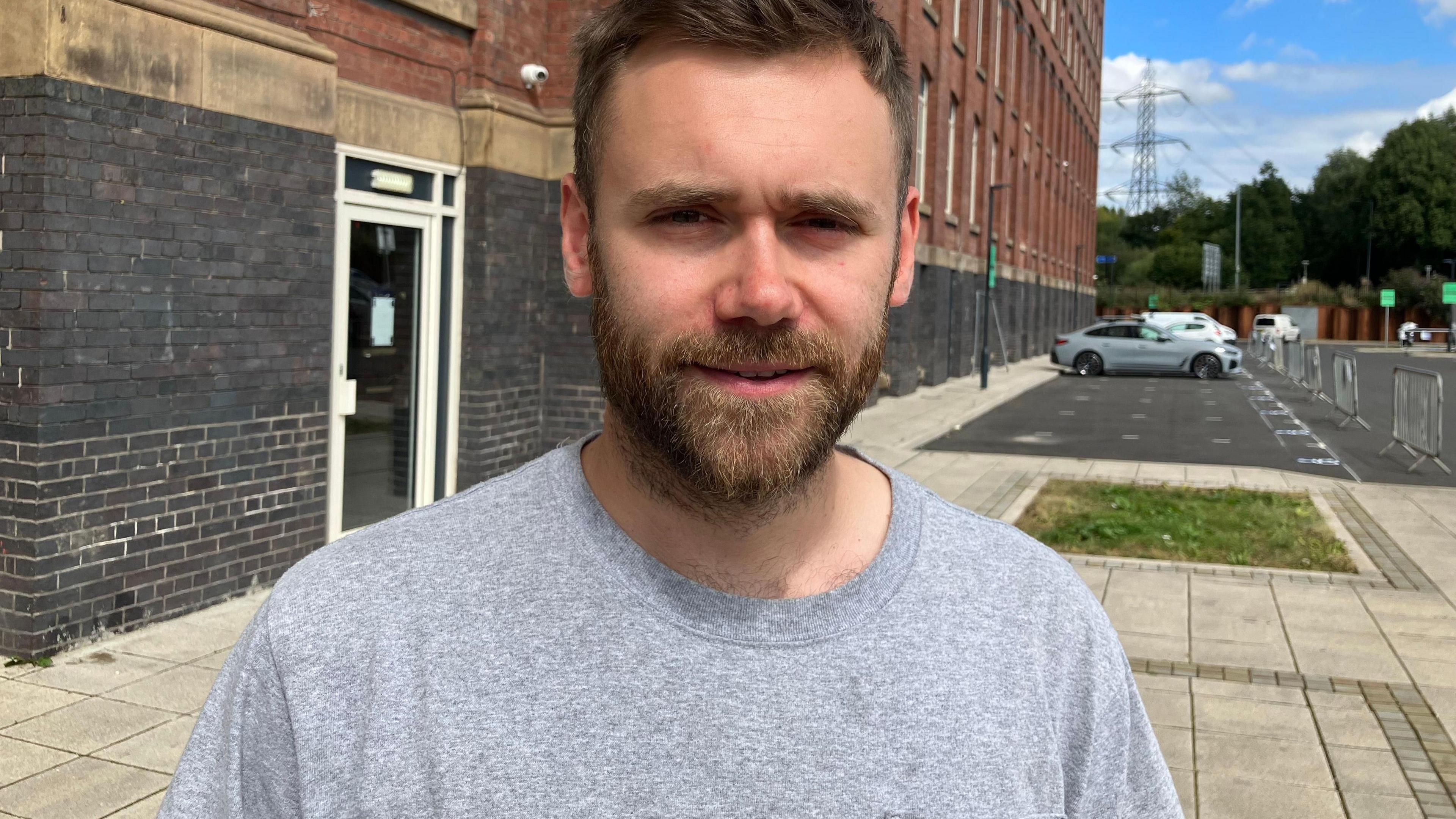 Doug Franks, who has brown hair and a brown beard and is wearing a grey t-shirt, smiles at the camera outside a converted mill building.