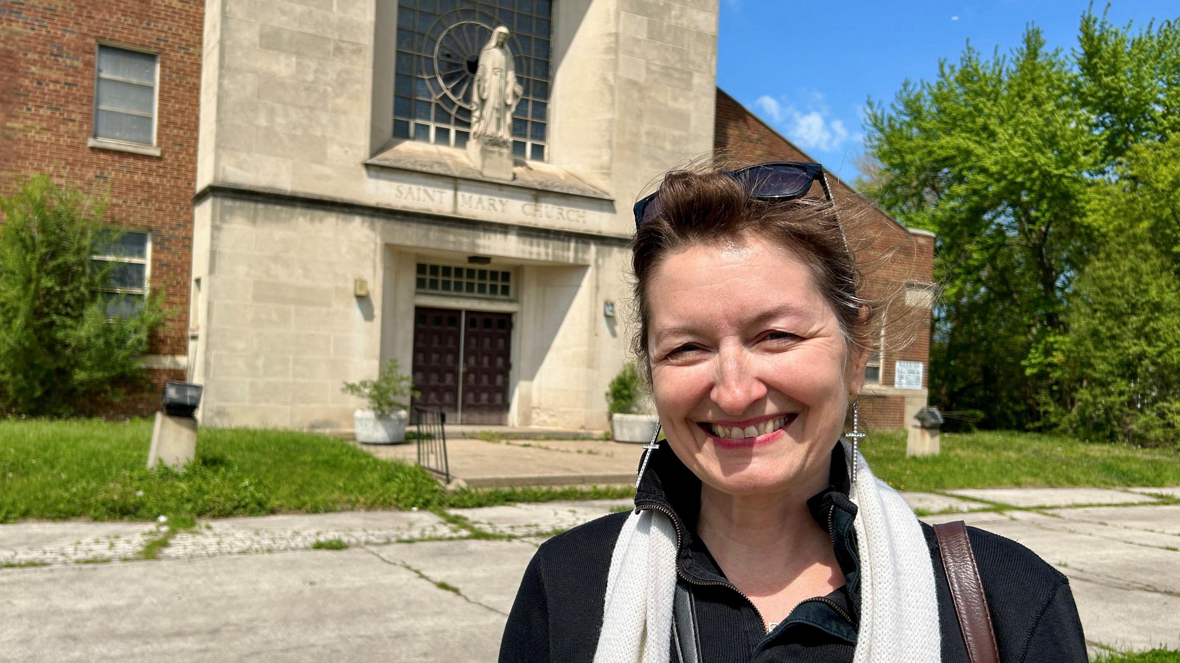 Mary Simons smiling outside of a stone fronted church with weeds in the background.
