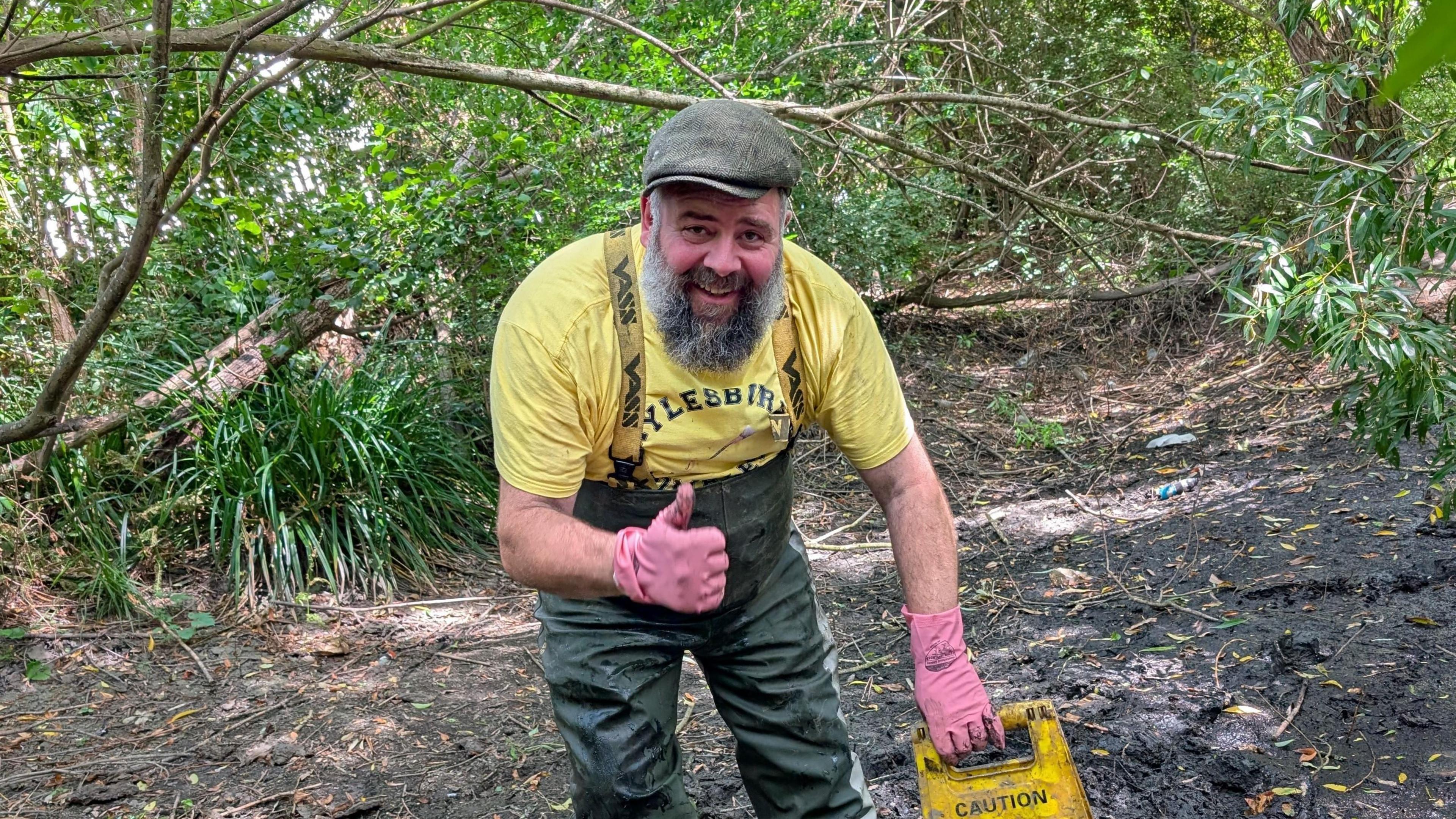 A smiling bearded man makes a thumbs up signs. He is wearing pink rubber gloves, waders and a yellow t-shirt. He is stood in a boggy river bed