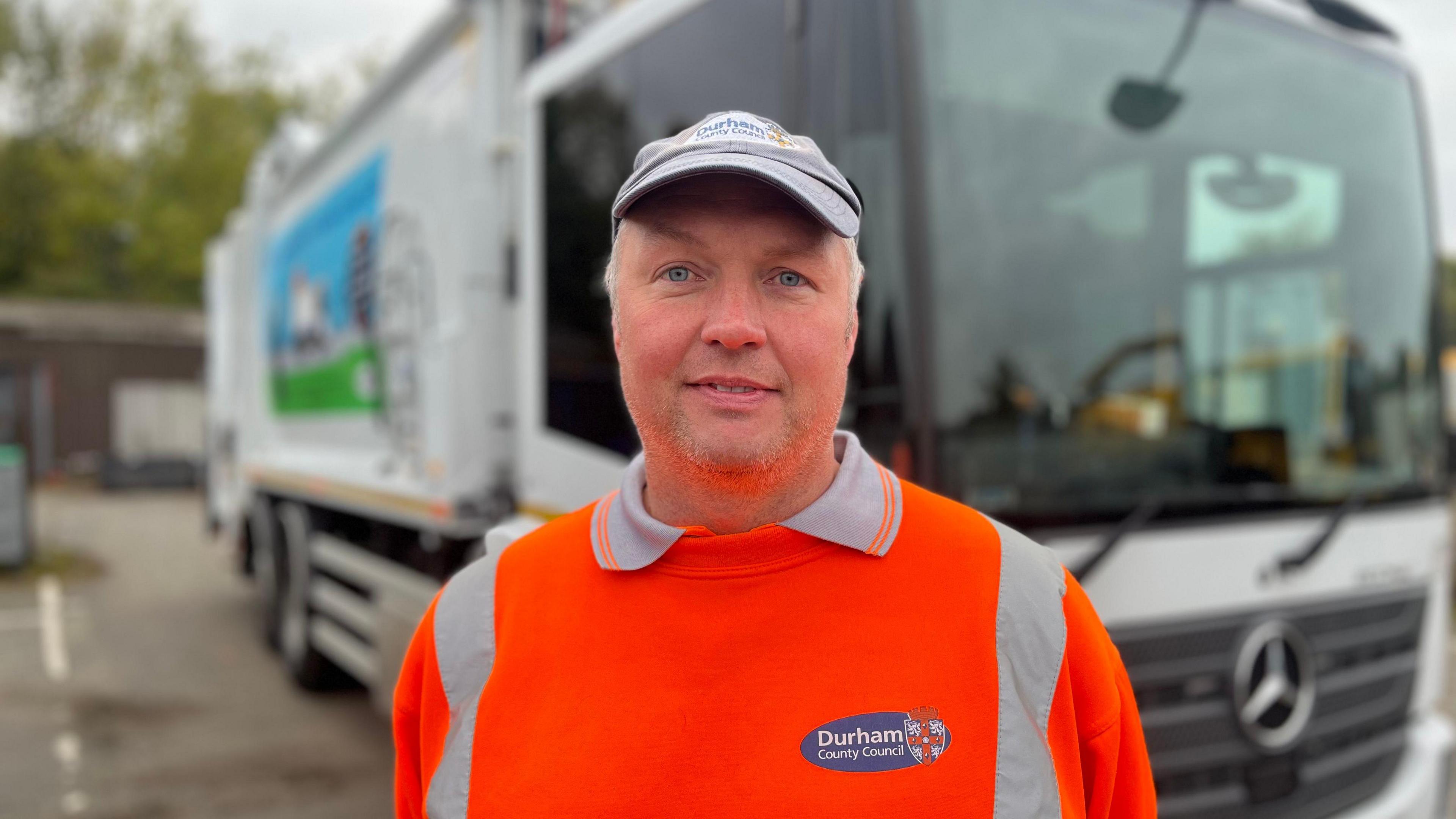 Craig Wilson wearing his orange Durham County Council uniform and a cap. He is standing in front of a bin lorry.