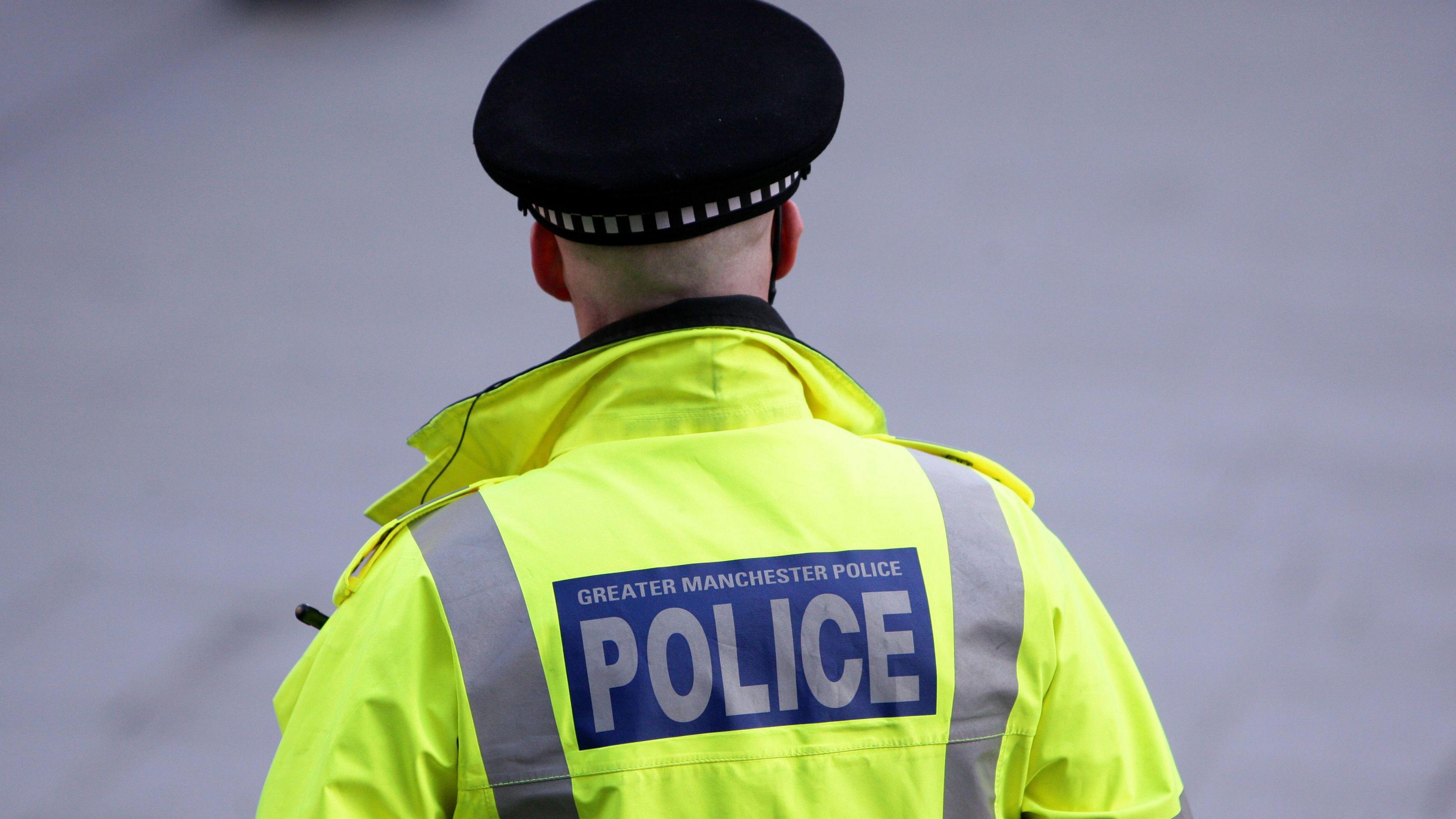 Generic image of the back of an unidentifiable policeman, who wears a black cap and yellow hi-vis jacket with blue logo saying Greater Manchester Police