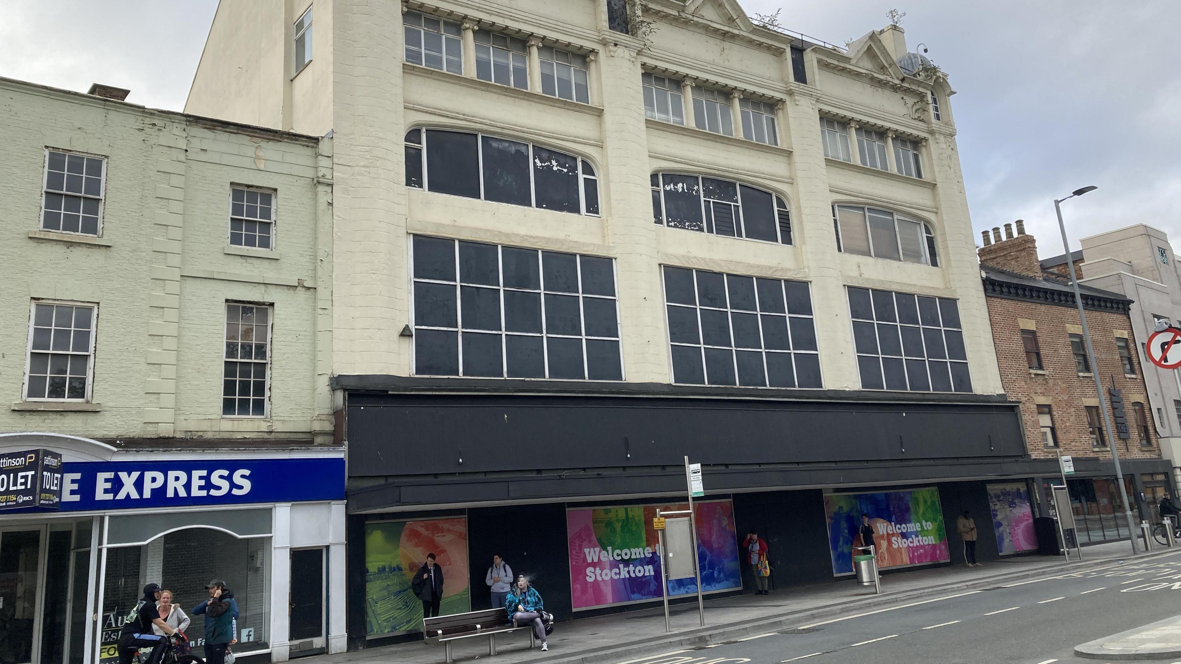 The former Debenhams department store in Stockton. It is a multi-storey building, painted cream with the majority of its windows blacked out. The lower area is painted black and is adorned with signs reading "Welcome to Stockton". Someone is sitting on a bench on the pavement, while three people stand chatting on the street nearby.