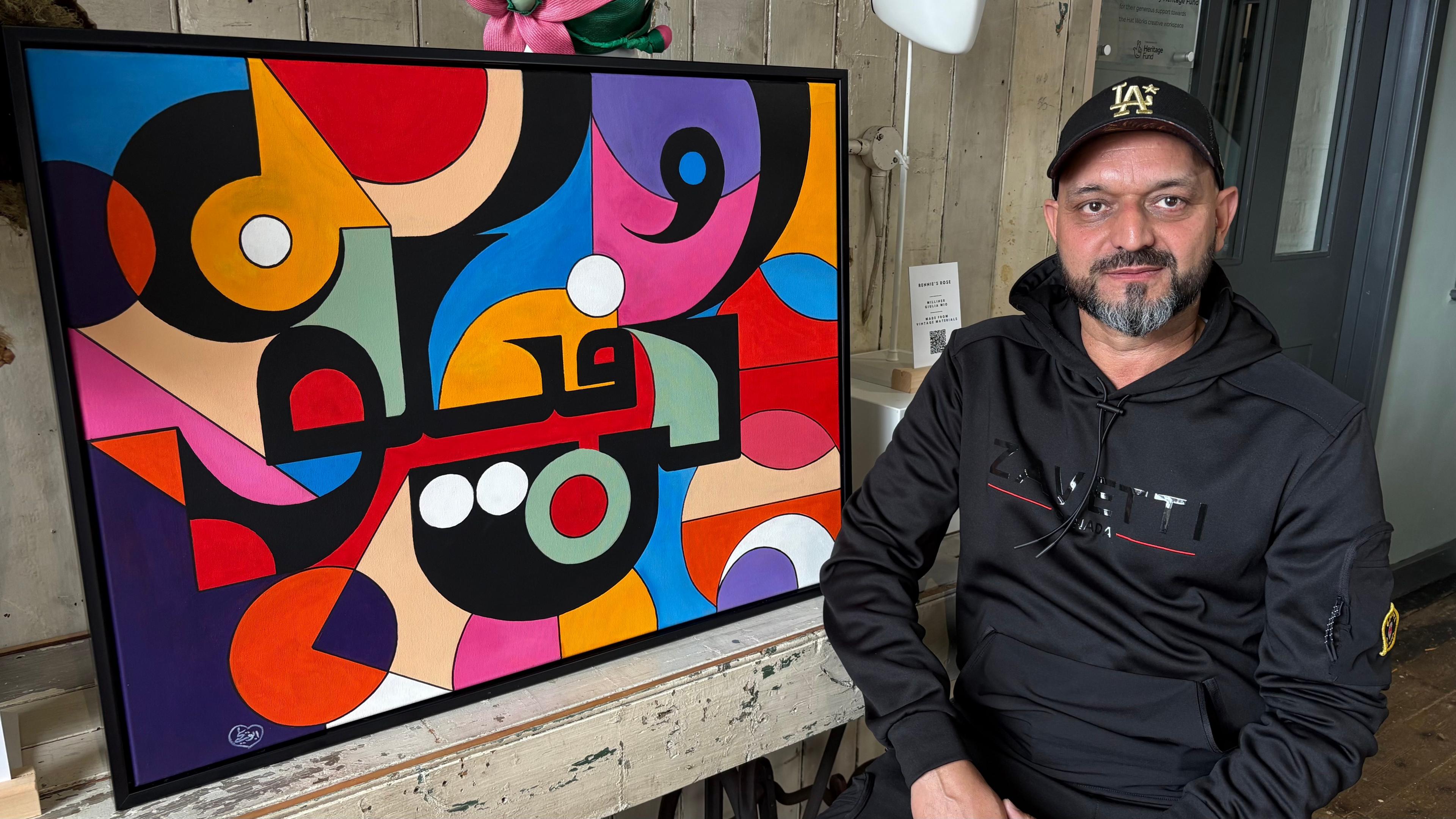 Abu-Yahya wearing a black cap and with a black beard, sitting next to an expressionist painting made up of richly-coloured shapes,