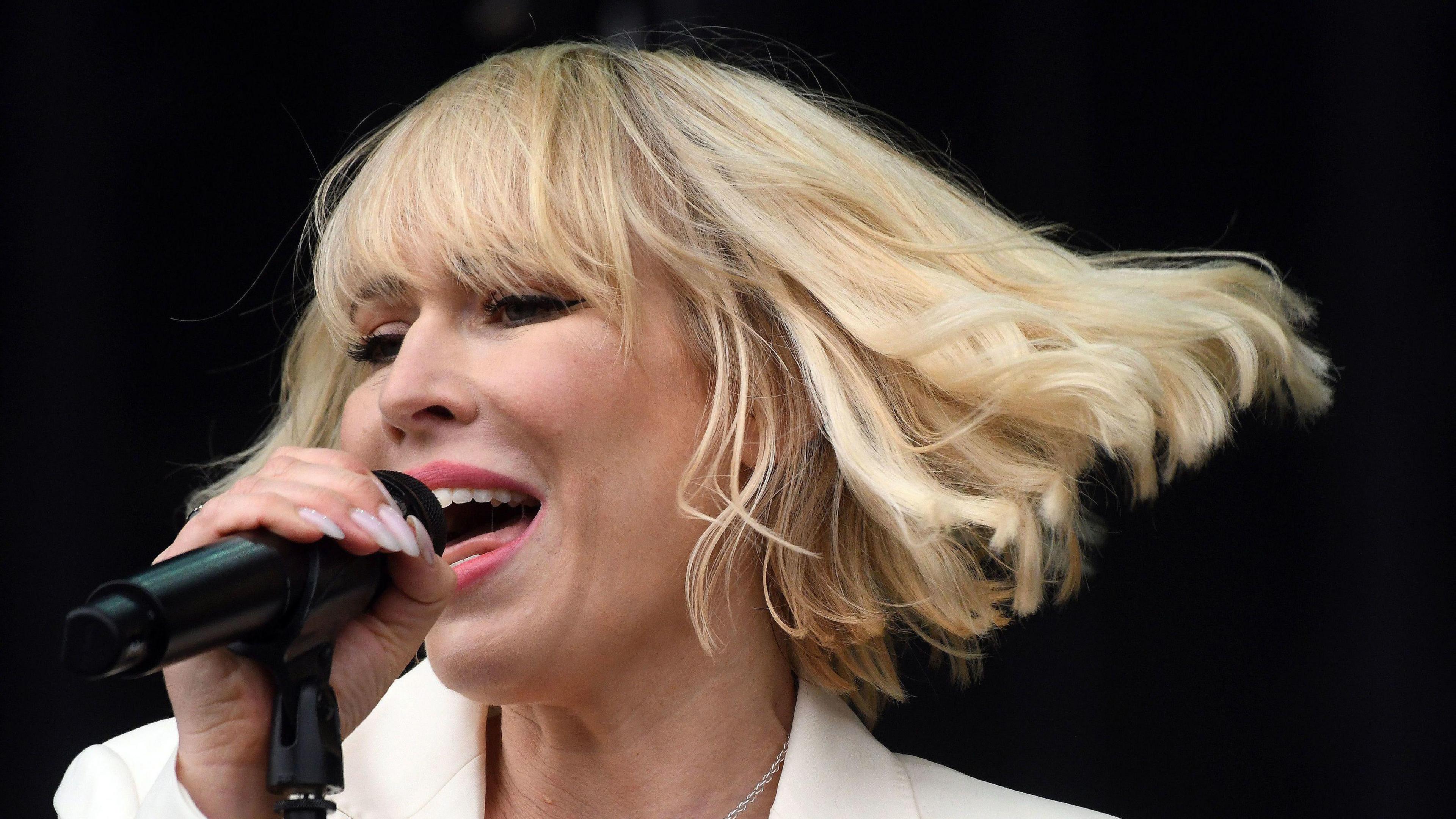 Natasha Bedingfield sings into a microphone, she has a blonde bob and is wearing a white suit jacket