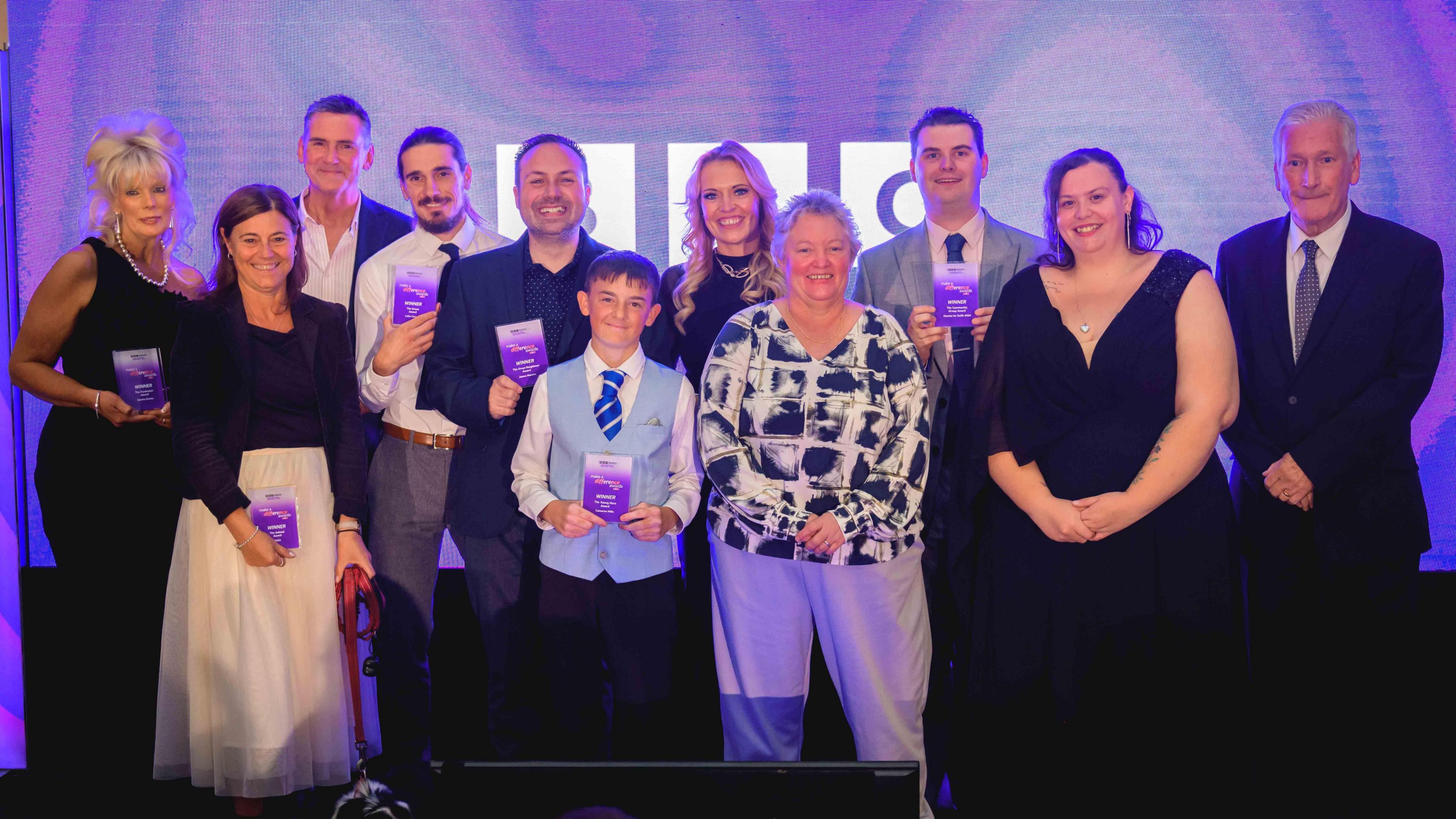 A group of people holding trophies stand on stage in front of a purple BBC background smiling at the camera.