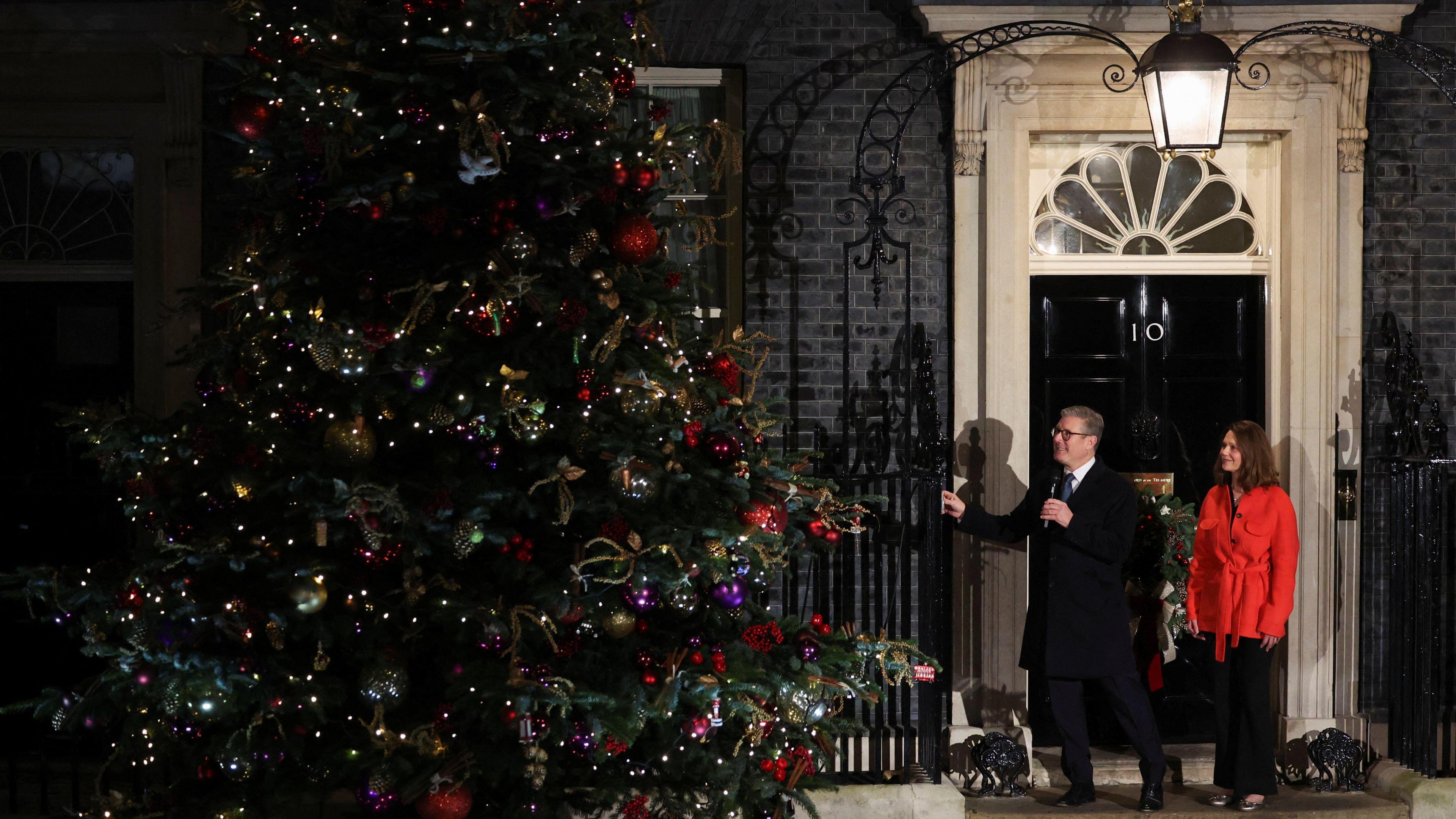 Sir Kier Starmer and his wife are standing at the door of 10 Downing Street. Sir Kier is pressing a button which turns on the Christmas lights. To their left is a large fir Christmas tree, covered in lights, as well as red, gold, silver and purple decorations.