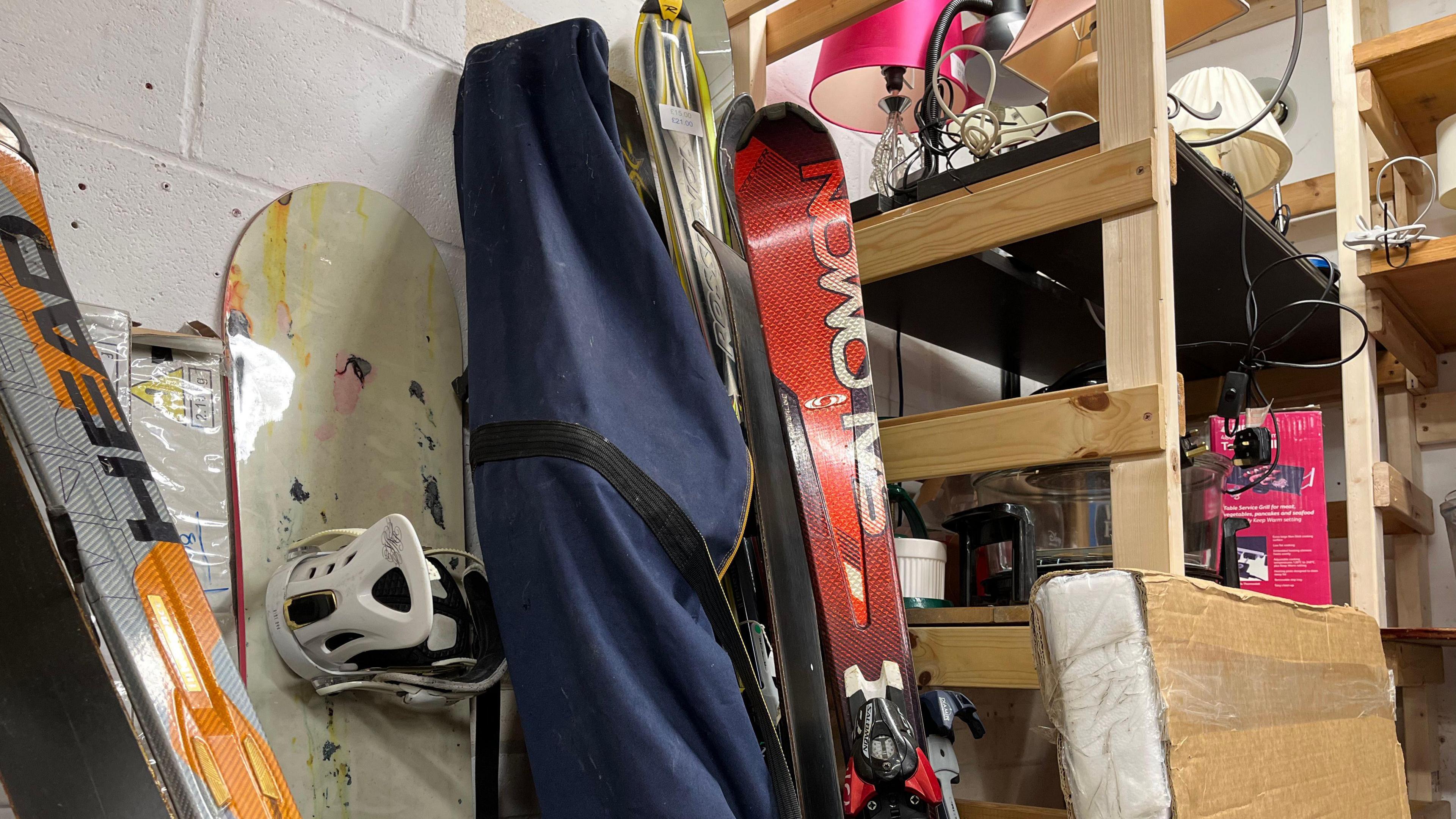 Skis and snowboards lean up against a wall, one set in a canvas bag, wooden shelves next to them full of lamps