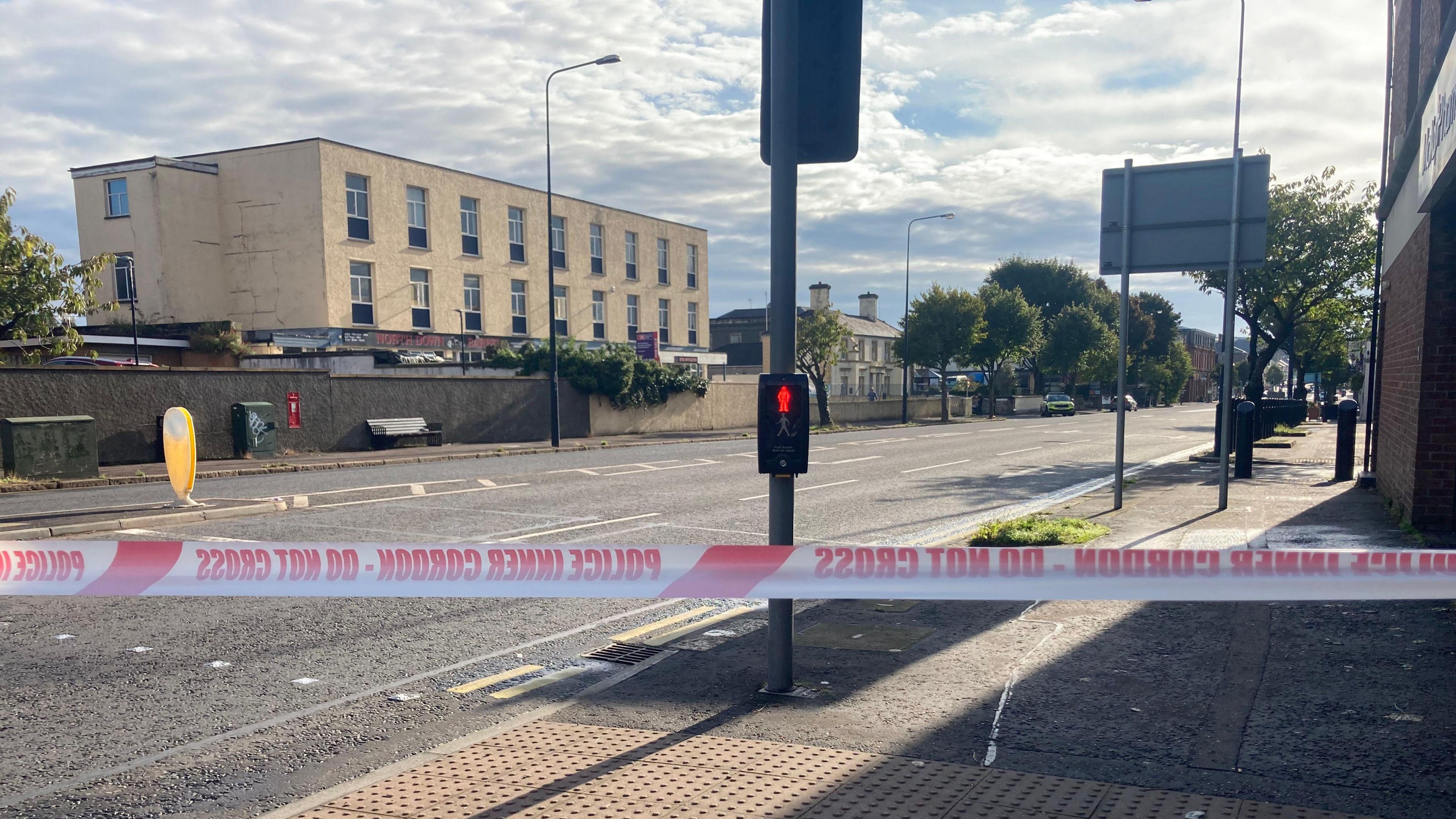 A street with a traffic light and crossing visible. There is a two-storey beige building to the left of the street. Police tape is across the centre of the image.